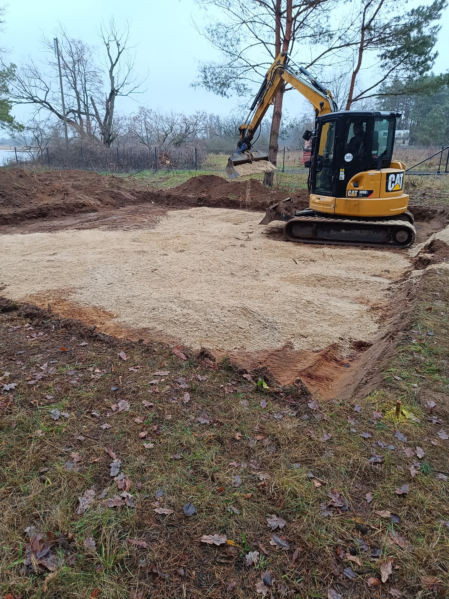 Żółta koparka CAT 305E2 wyrównuje teren pod budowę fundamentów, widoczne wykopane obrysy, przygotowane podłoże z piasku, w tle drzewa bez liści i fragment jeziora.