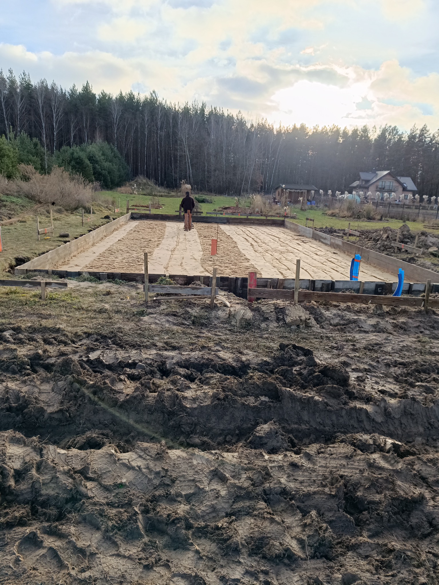 Przygotowanie terenu pod wylanie fundamentów: szalunek z drewnianych desek, piasek wewnątrz, widoczne rury instalacyjne i pracownik zacierający piasek, w tle las i dom.