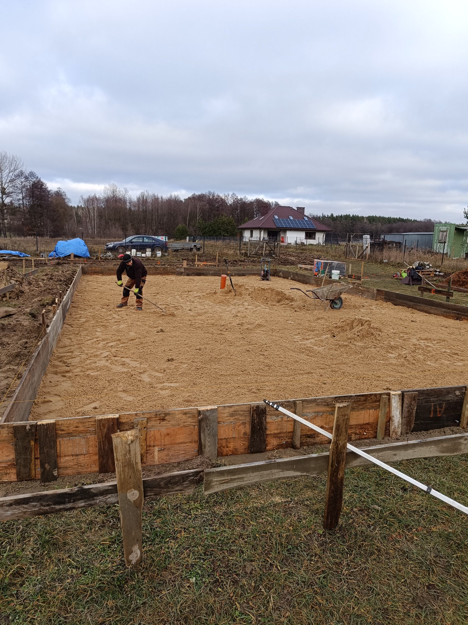 Przygotowanie podłoża pod fundament: pracownik wyrównuje piasek w drewnianym szalunku, w tle widoczny dom z panelami słonecznymi.