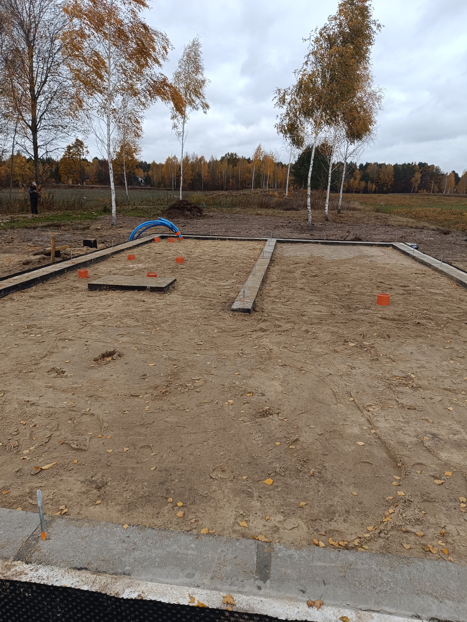 Przygotowany teren pod fundamenty z widocznymi obrysami ścian i słupów, pomarańczowe rury instalacyjne i jesienne drzewa w tle.