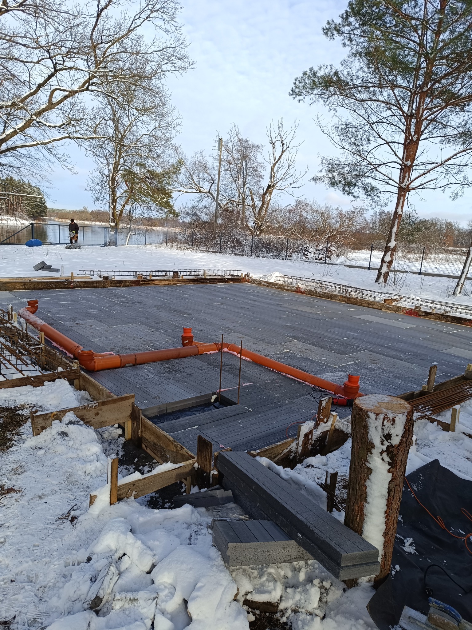 Przygotowany teren pod wylewkę fundamentów w zimowej scenerii, widoczne zbrojenie, rury kanalizacyjne i drewniane szalunki, w tle zalesiony brzeg jeziora.