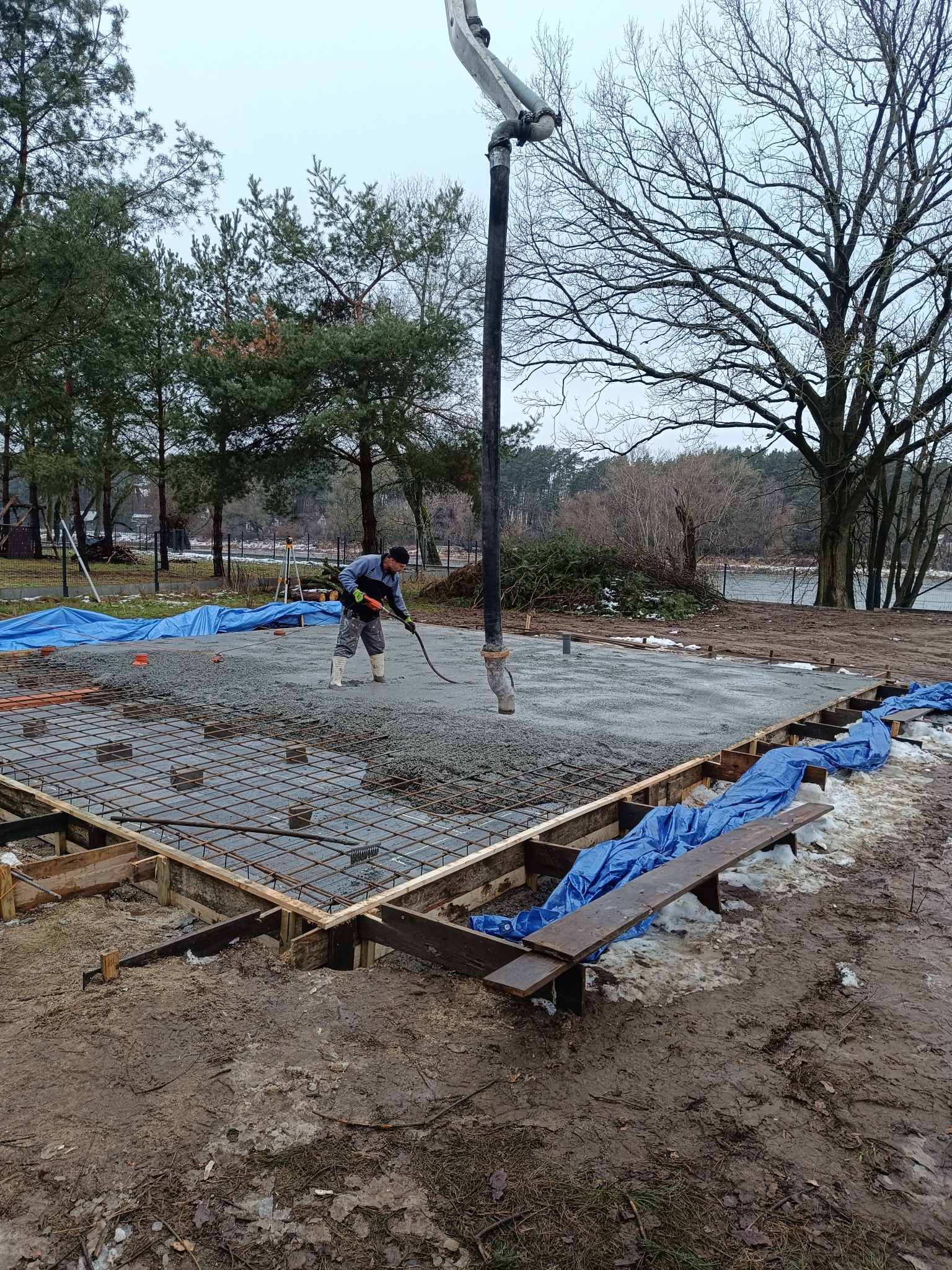 Wylewanie betonu na przygotowany zbrojony fundament pod budowę domu jednorodzinnego, widoczny pracownik wyrównujący beton, rura do podawania betonu, szalunek i siatka zbrojeniowa, w tle drzewa...