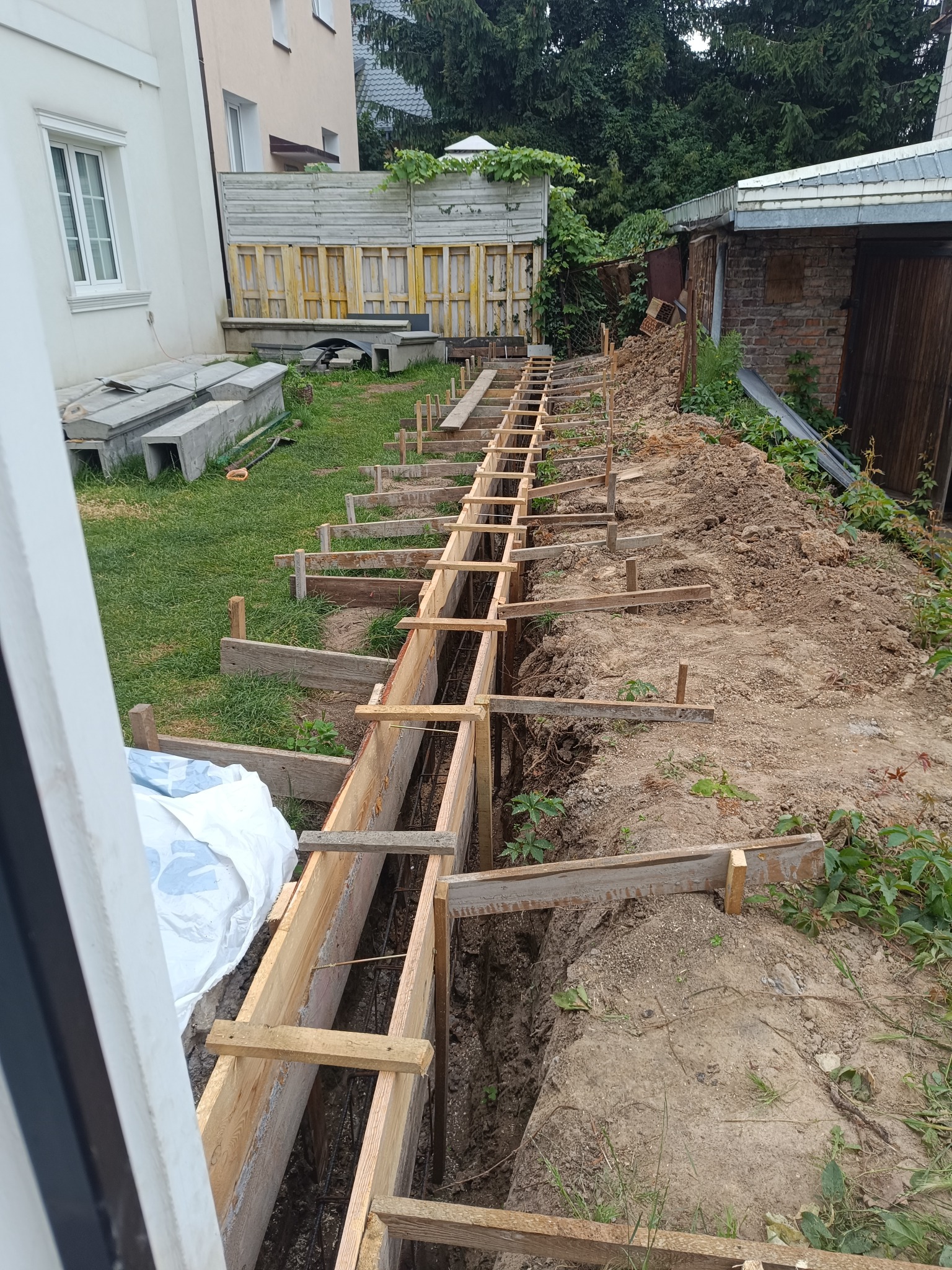 Długi wykop z drewnianym szalunkiem i zbrojeniem, przygotowany pod wylanie fundamentów. Widoczne elementy konstrukcyjne i otoczenie placu budowy.