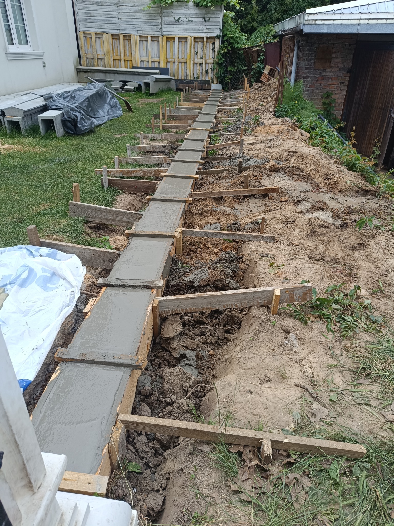 Wylany beton w szalunku z desek, tworzący fundament pod ogrodzenie na nierównym terenie. Widoczne elementy konstrukcji i otoczenie budowy.