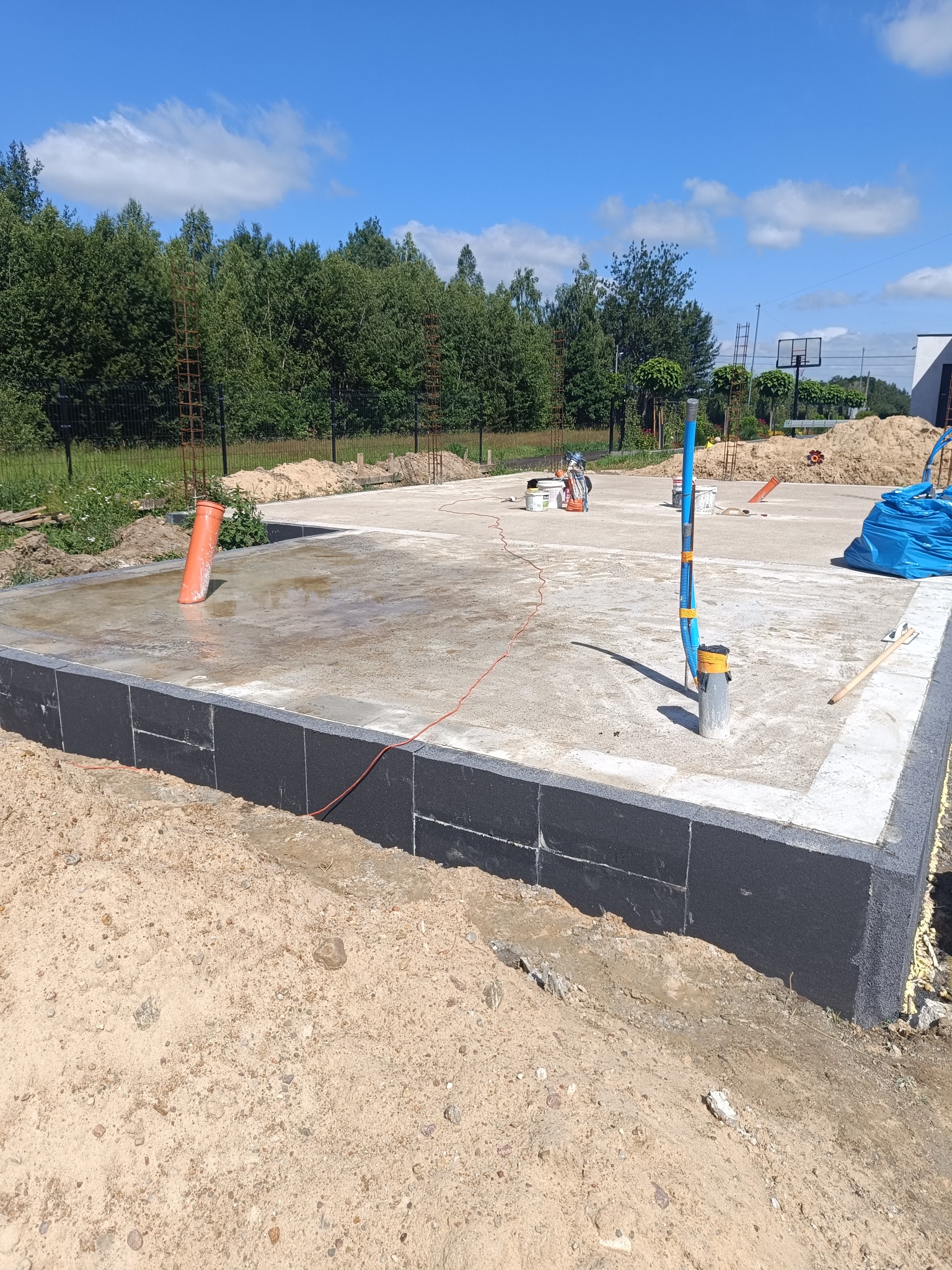 Zalany fundament z wystającymi rurami instalacyjnymi i czarną izolacją pionową, w tle drzewa i błękitne niebo. Widoczne narzędzia i materiały budowlane.