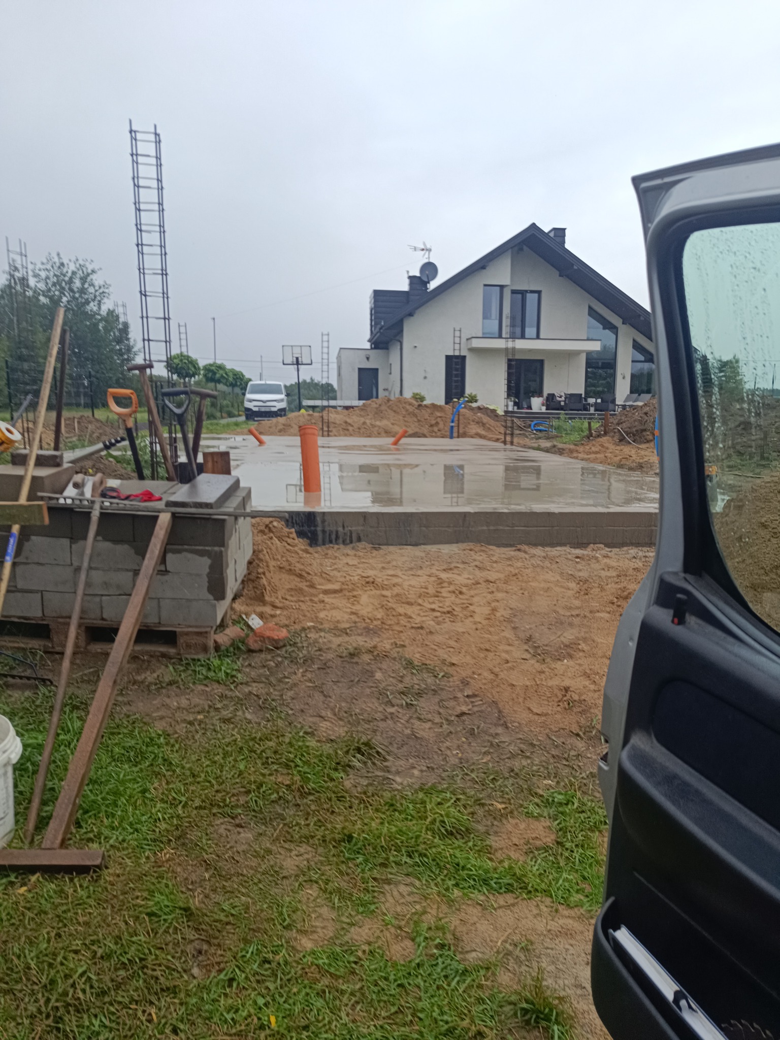 Wylany fundament pod dom jednorodzinny z widocznymi narzędziami i częściowo otwartymi drzwiami samochodu w pochmurny dzień. W tle biały dom z ciemnym dachem.