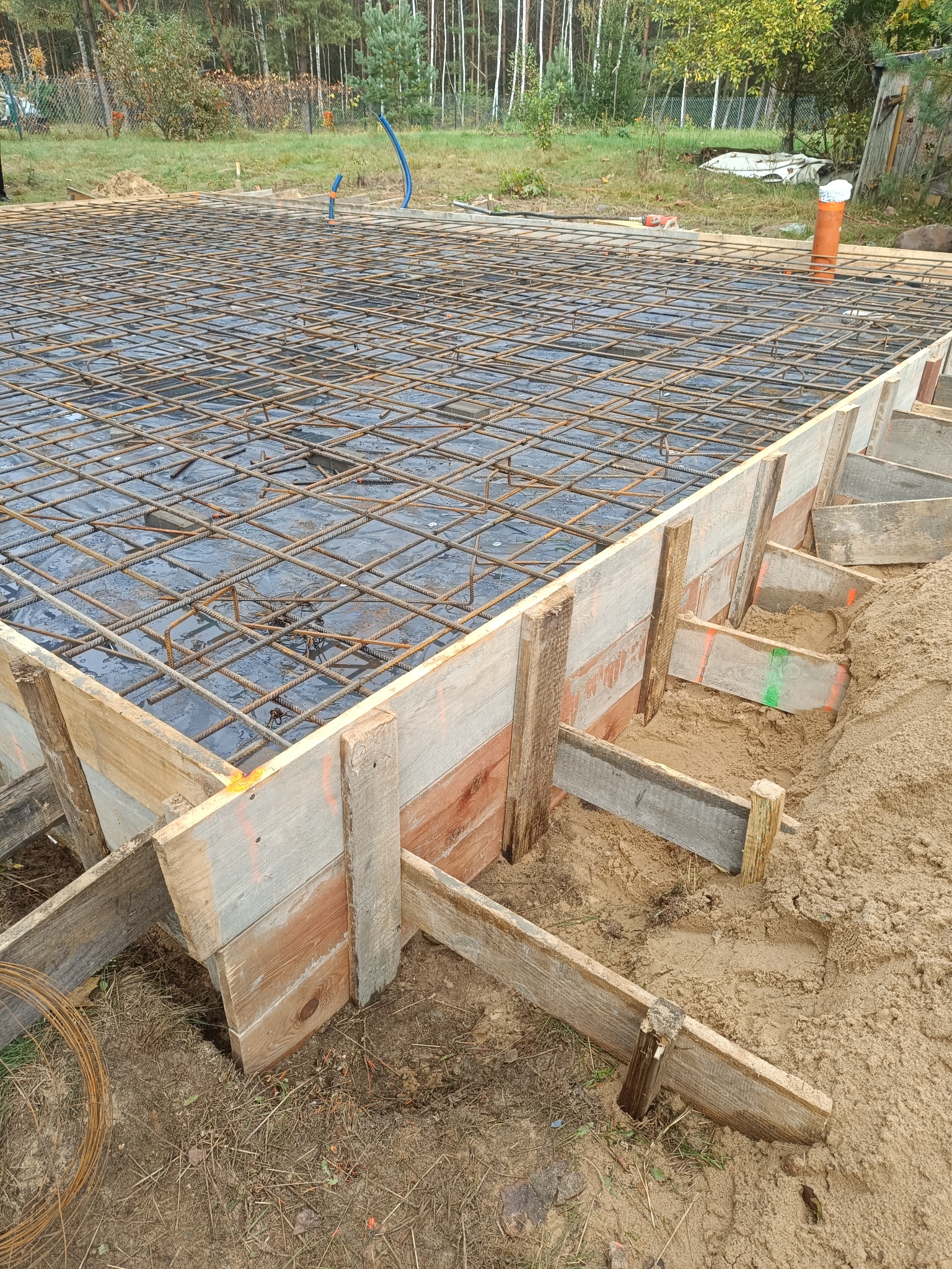 Zbrojenie fundamentów w drewnianym szalunku, widoczne pręty i folia. Prace ziemne, teren budowy z piaskiem i zielenią w tle. Elementy konstrukcyjne z bliska.