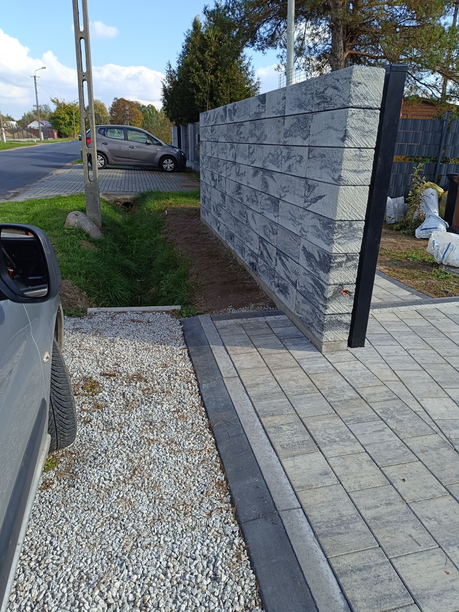 Nowoczesny, szary murek z betonowych bloczków, pełniący funkcję ogrodzenia, z widoczną kostką brukową i fragmentem parkingu żwirowego. Słoneczny dzień.