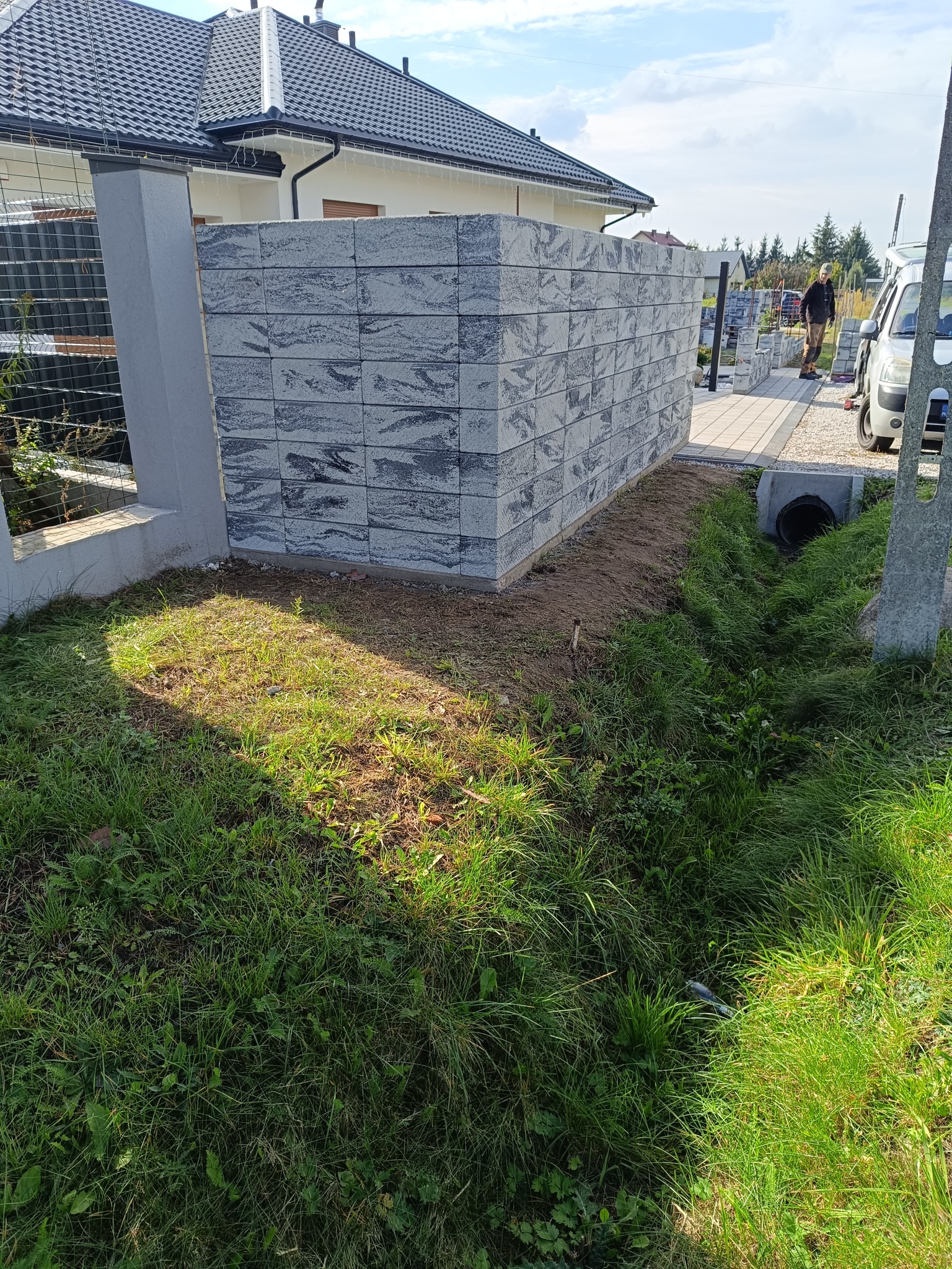 Nowoczesny, szary murek z bloczków betonowych, częściowo ukończony, z widocznym fundamentem, na tle domu z ciemną dachówką i pracownikiem w oddali.