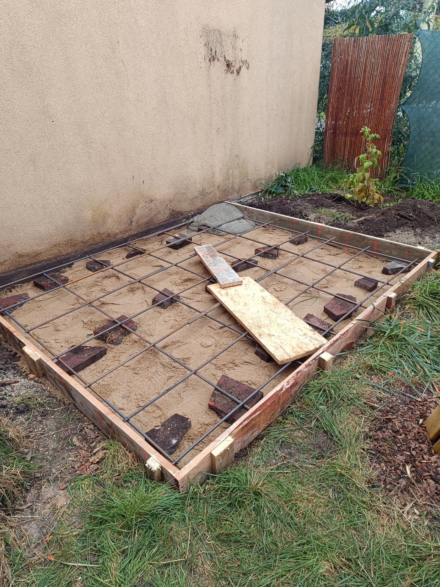 Szablon z desek, siatka zbrojeniowa i cegły na piasku, przygotowane pod wylewkę betonową fundamentu. Widok z góry na plac budowy.