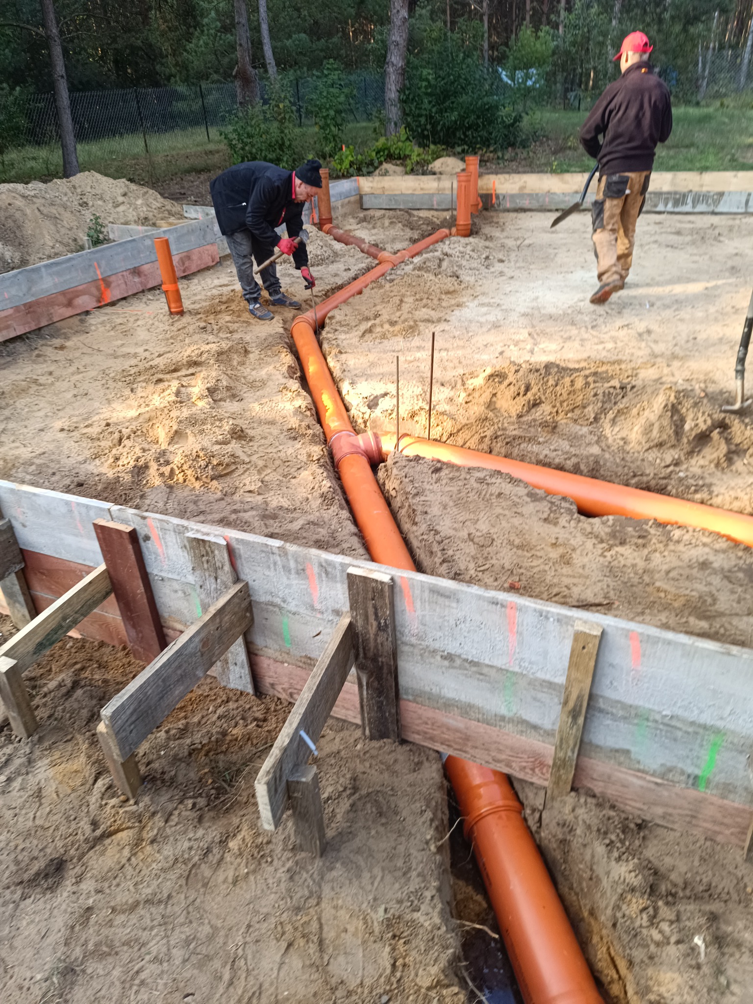 Fundamenty: montaż rur kanalizacyjnych w wykopie z szalunkiem. Dwóch pracowników przy pracy, jeden zagęszcza ziemię, drugi z łopatą. Pomarańczowe rury.