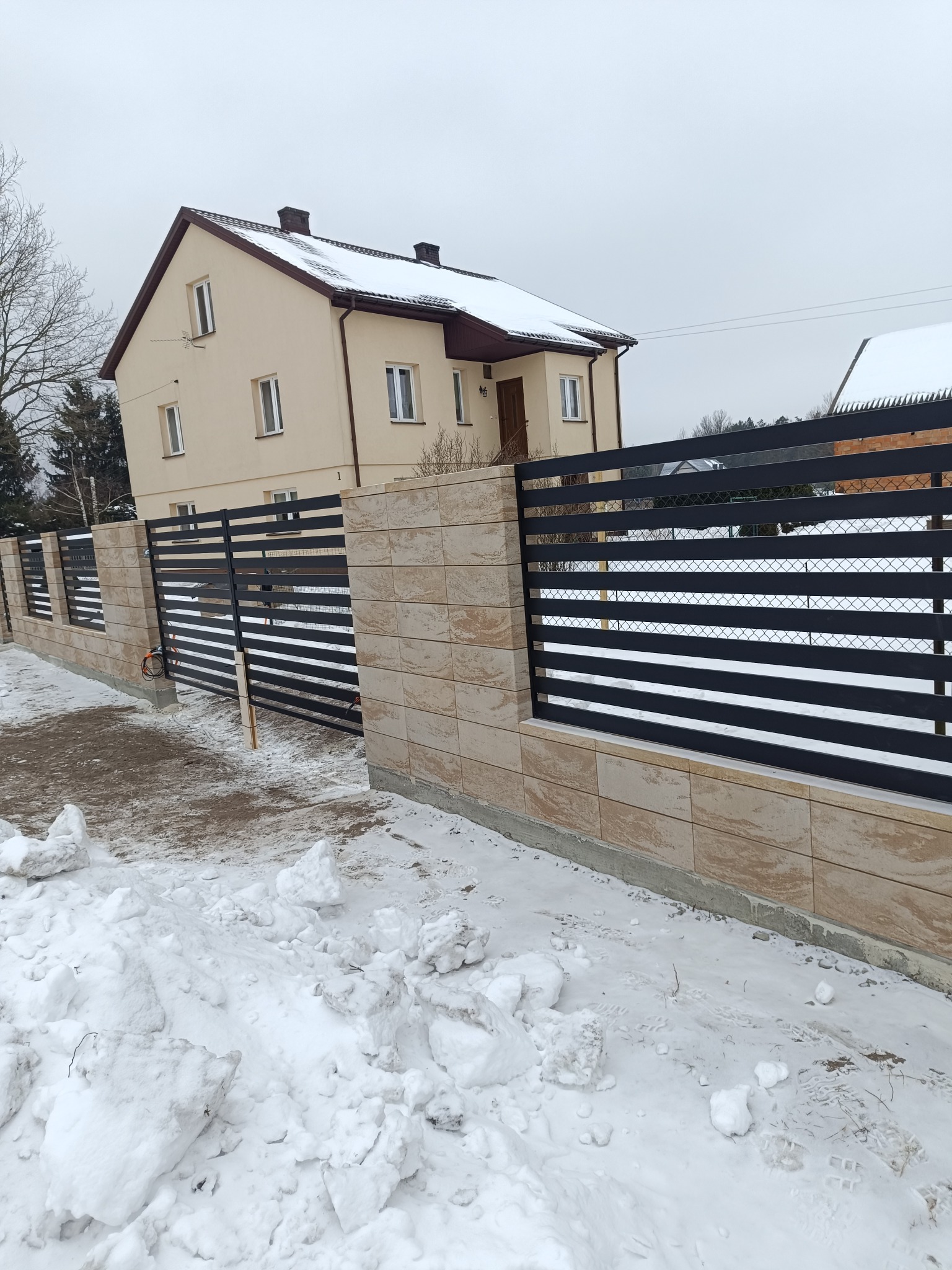 Nowoczesne ogrodzenie panelowe z elementami z płytek imitujących kamień, w zimowej scenerii. Dom w tle.