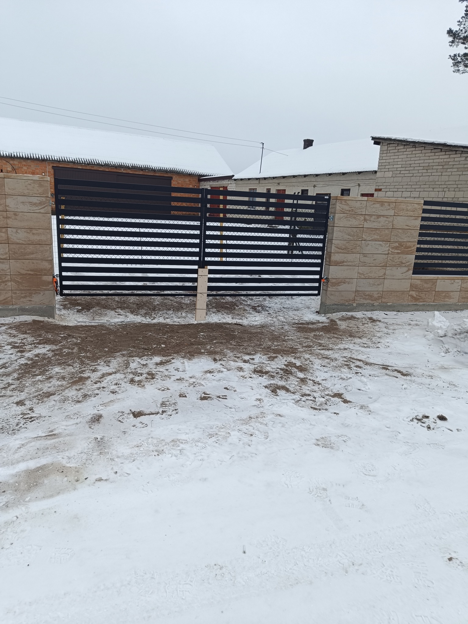 Nowoczesna, pozioma brama wjazdowa z czarnymi przęsłami i beżowymi słupkami z płytek, na tle zimowego krajobrazu z budynkami pokrytymi śniegiem.