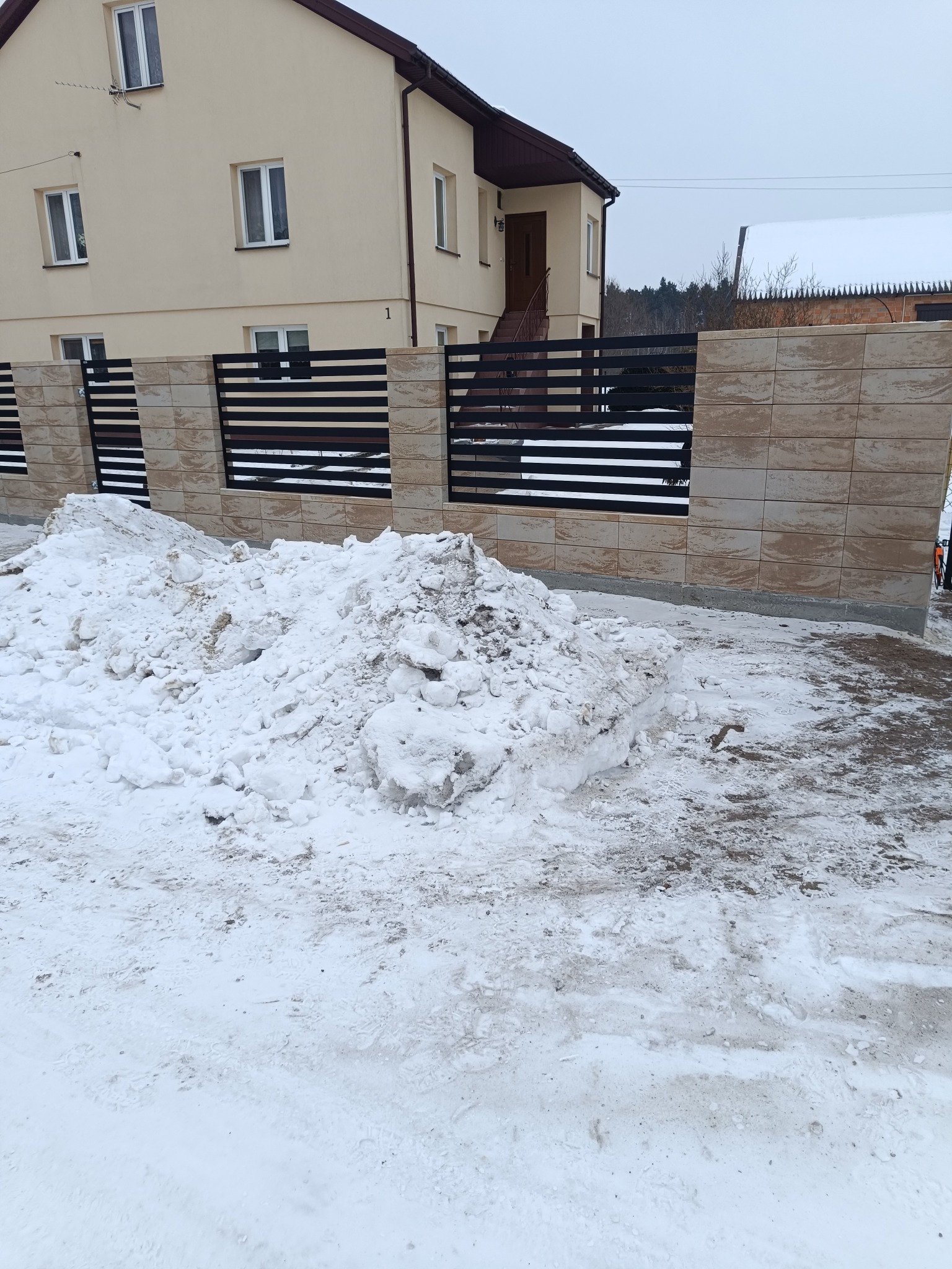 Nowoczesne ogrodzenie panelowe z elementami z piaskowca, widok z zewnątrz w zimowej scenerii. Dom z beżową elewacją w tle.