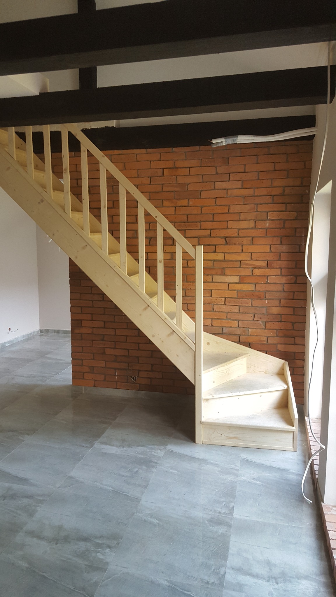 Surowe drewniane schody z balustradą na tle ceglanej ściany i szarej podłogi z płytek imitujących kamień, widoczne czarne belki stropowe.