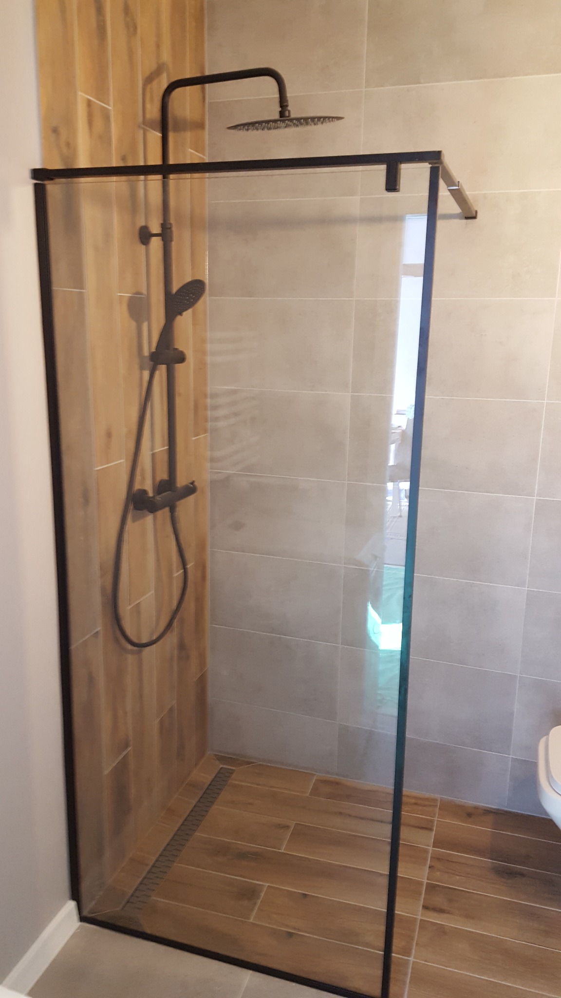 Nowoczesna kabina prysznicowa typu walk-in z czarną armaturą i odpływem liniowym, wykończona płytkami imitującymi drewno i szarymi płytkami.