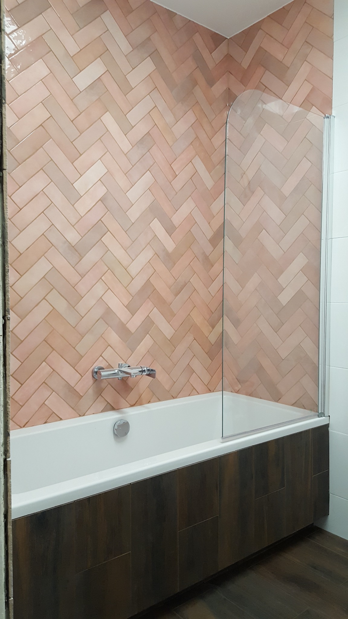 Łazienka z wanną obudowaną ciemnym drewnem i ścianą wyłożoną różową cegiełką ułożoną w jodełkę, z zamontowanym szklanym parawanem nawannowym i chromowaną armaturą.