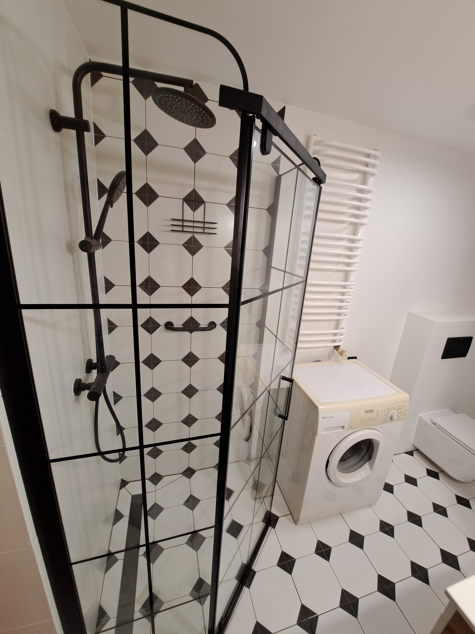 Nowoczesna łazienka z czarną kabiną prysznicową typu walk-in, białą pralką i grzejnikiem drabinkowym, wykończona biało-czarną mozaiką podłogową.