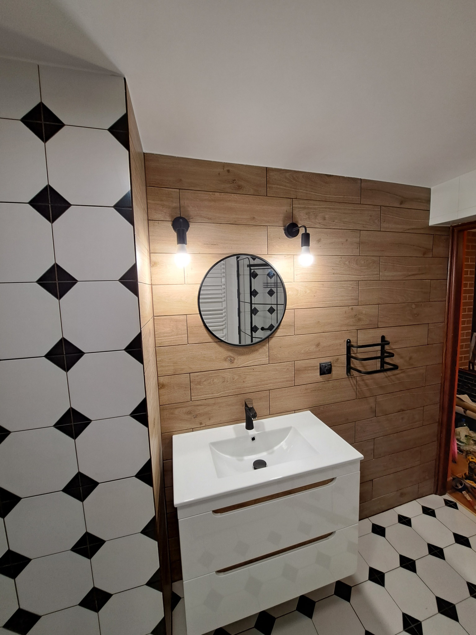 Nowoczesna łazienka z drewnopodobnymi płytkami ściennymi, białą szafką z umywalką, okrągłym lustrem i czarno-białymi płytkami podłogowymi w romby.