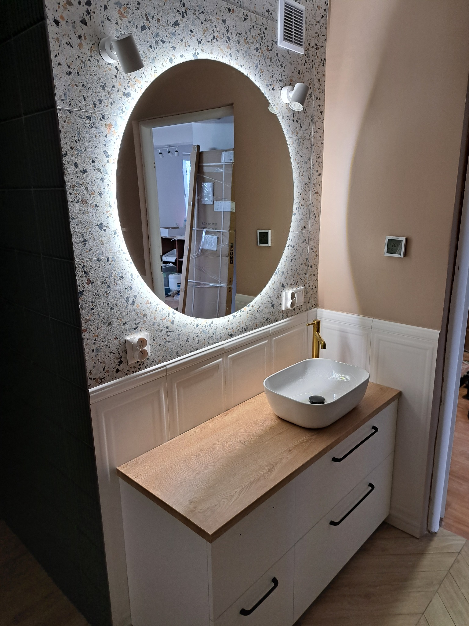 Nowoczesna łazienka z owalnym lustrem podświetlanym LED, umywalką nablatową na szafce z drewnianym blatem i złotą baterią, ściany wykończone płytkami lastryko i panelami ściennymi.