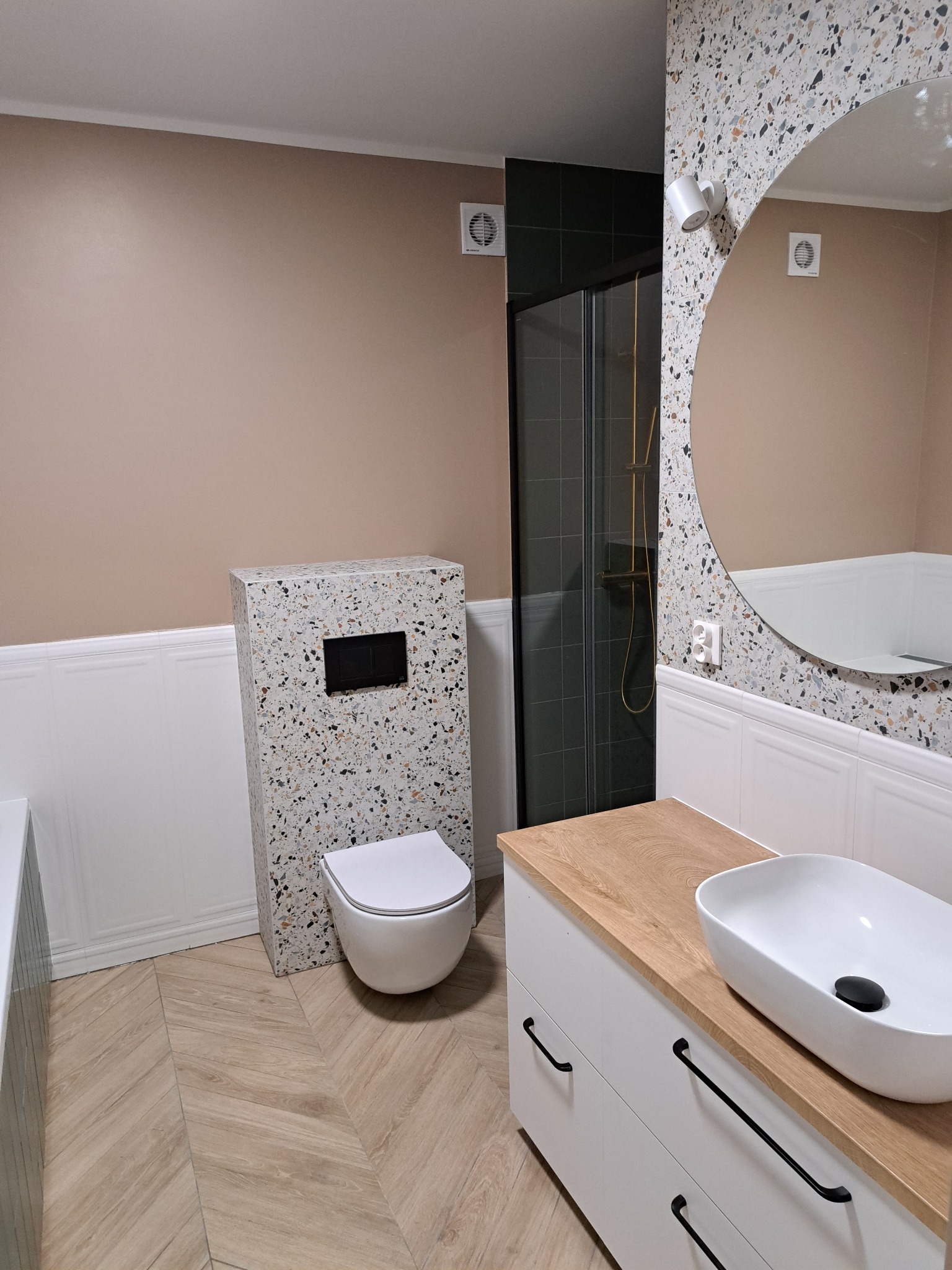 Nowoczesna łazienka z płytkami lastryko na obudowie geberitu i ścianie, jasne drewnopodobne płytki na podłodze ułożone w jodełkę, biała szafka pod umywalką z drewnianym blatem i czarnymi uchwytami,...