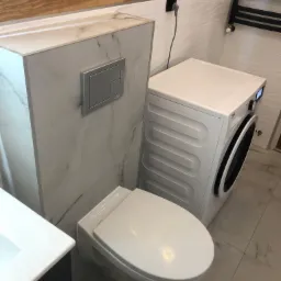 Nowoczesna łazienka z podwieszaną miską WC obudowaną płytą imitującą marmur, pralką Beko i grzejnikiem drabinkowym na ścianie wykończonej płytkami z delikatnym wzorem fal.