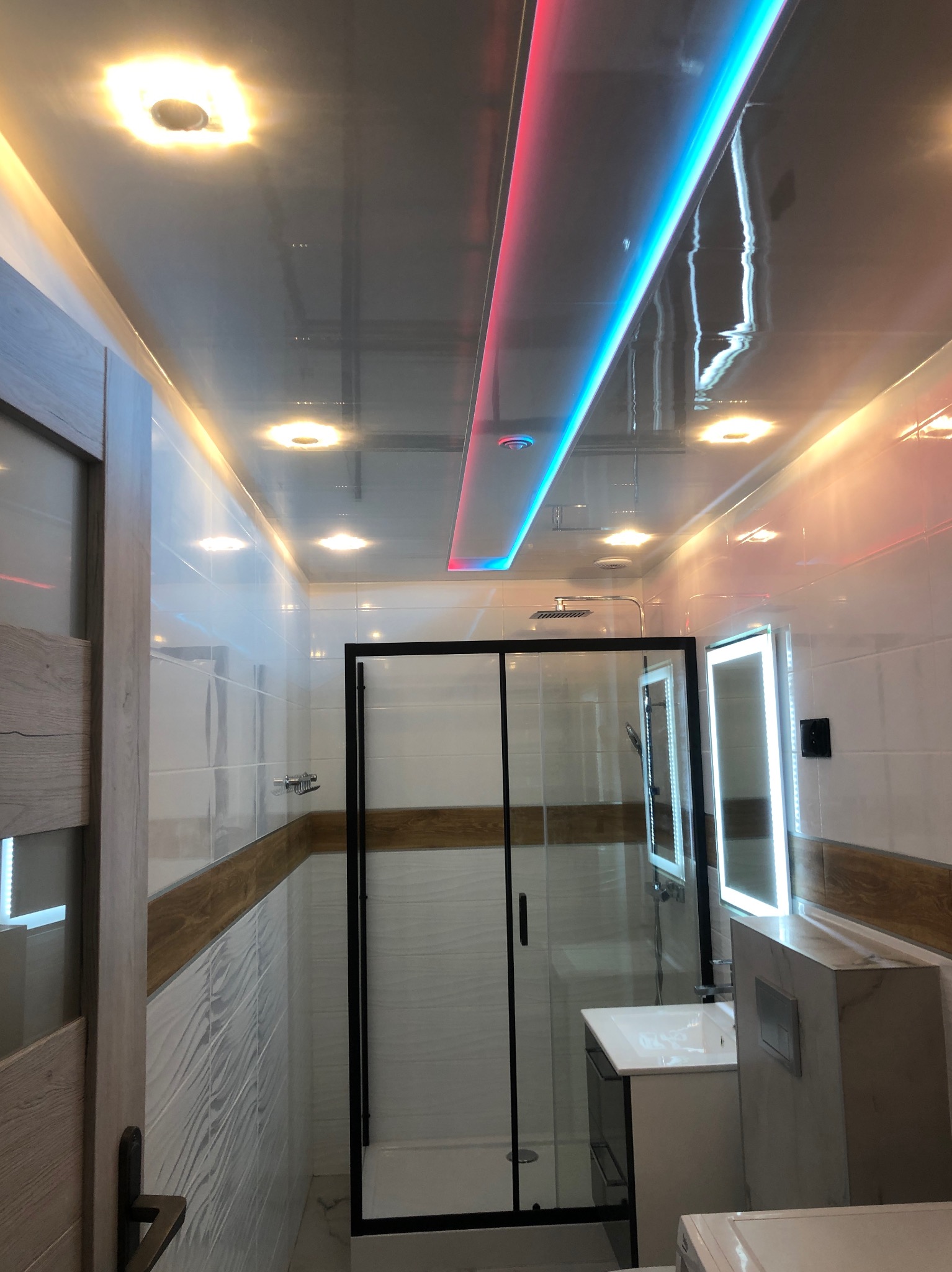 Nowoczesna łazienka z błyszczącym sufitem podwieszanym, oświetleniem LED w kolorach niebieskim i czerwonym, kabiną prysznicową z czarną ramą, podświetlanym lustrem i umywalką z marmurowym blatem.