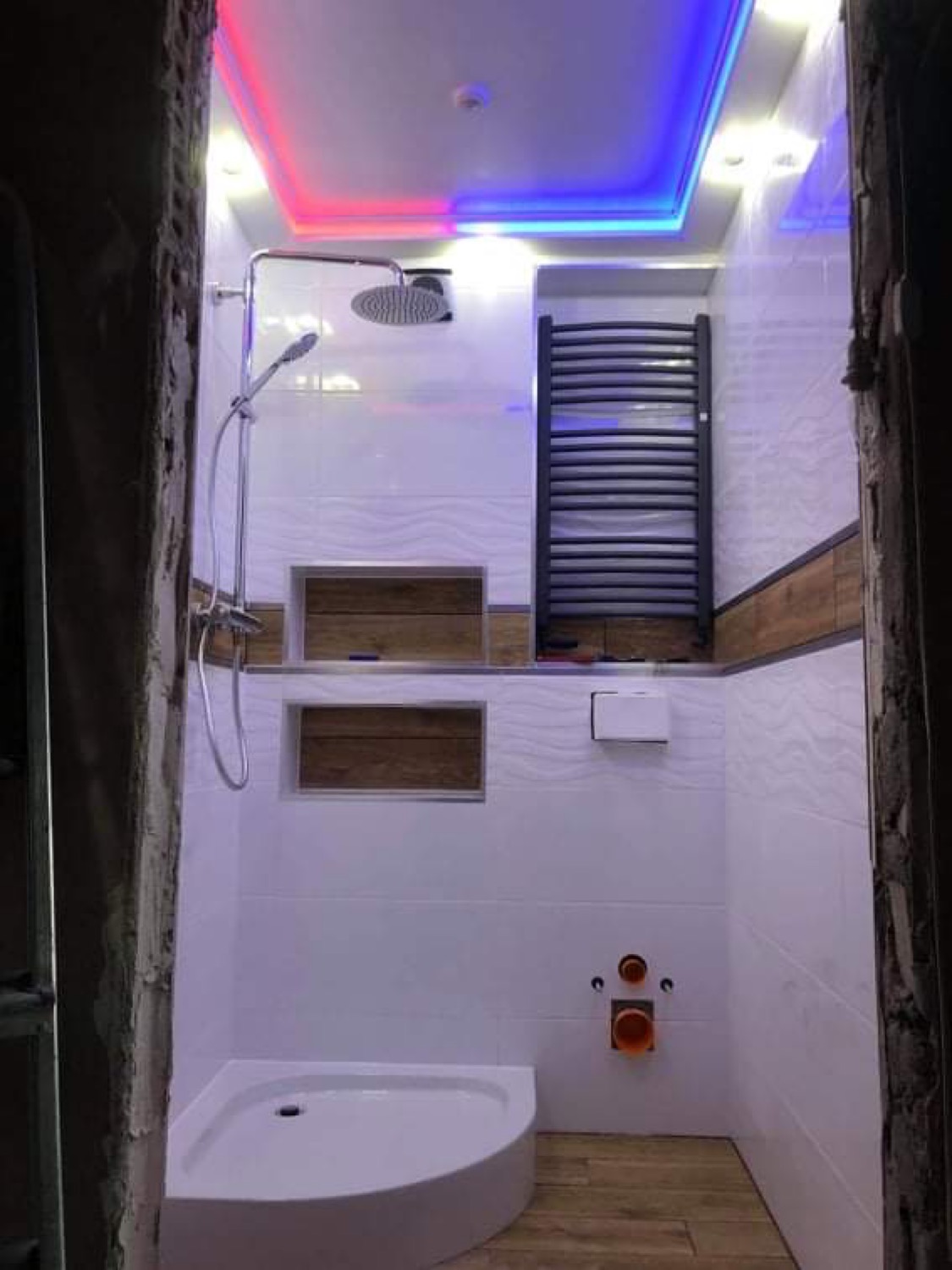 Nowoczesna łazienka po remoncie z kabiną prysznicową, grzejnikiem drabinkowym i podświetleniem LED w kolorach czerwonym i niebieskim, widok zza futryny