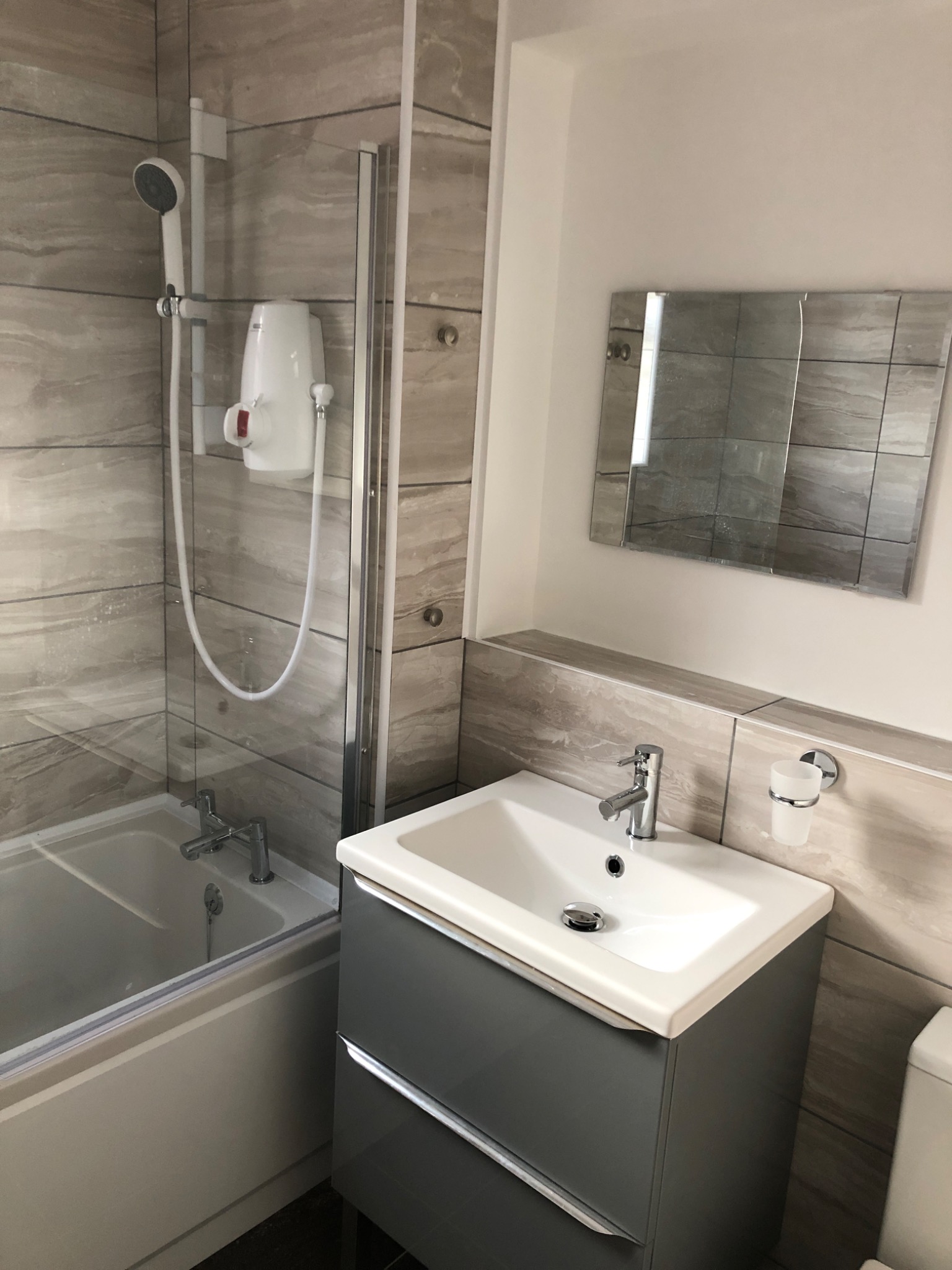 Wykończona łazienka z wanną z parawanem, umywalką z szarą szafką, lustrem z płytek i beżowymi płytkami imitującymi drewno na ścianach.