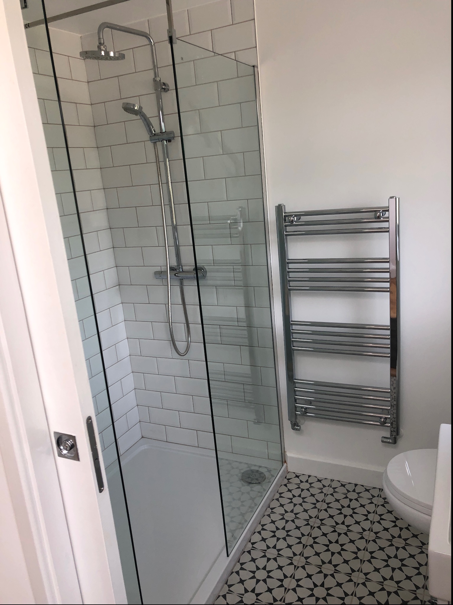 Nowoczesna kabina prysznicowa z białymi, prostokątnymi płytkami, chromowaną armaturą i grzejnikiem drabinkowym na tle wzorzystej podłogi w łazience.