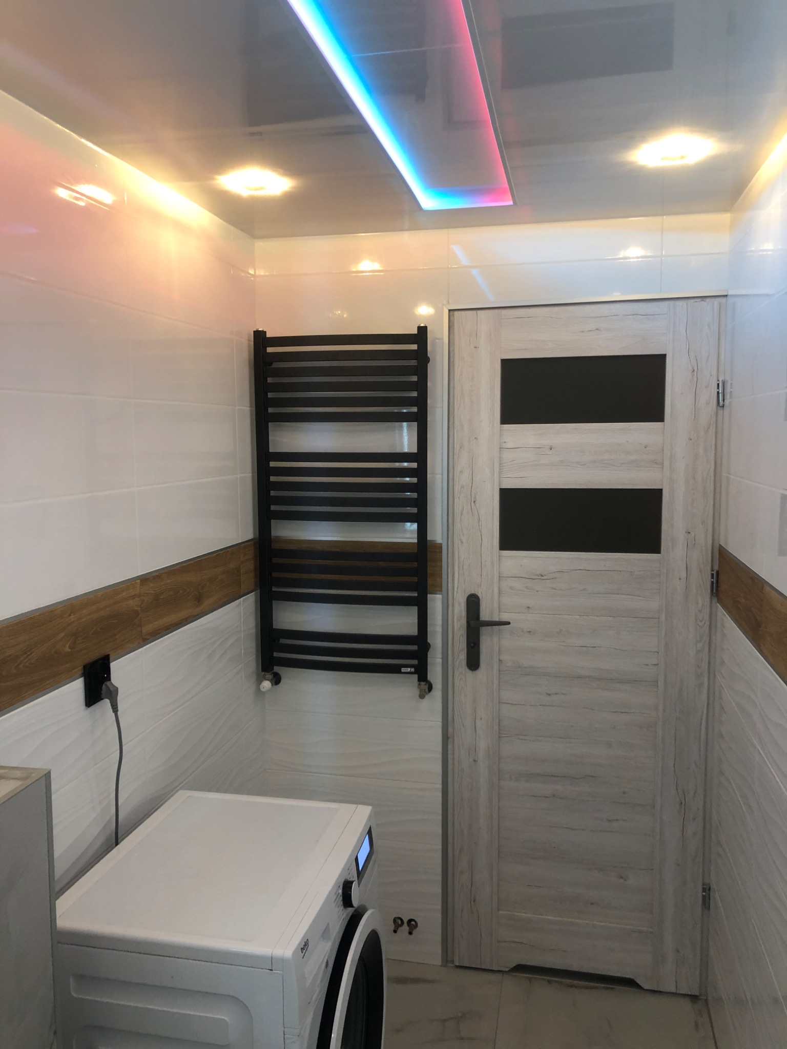 Wykończona łazienka z biało-szarymi płytkami imitującymi fale, drewnopodobnym pasem dekoracyjnym, grzejnikiem drabinkowym, pralką Beko oraz drzwiami z drewnopodobną okleiną i czarnymi wstawkami...