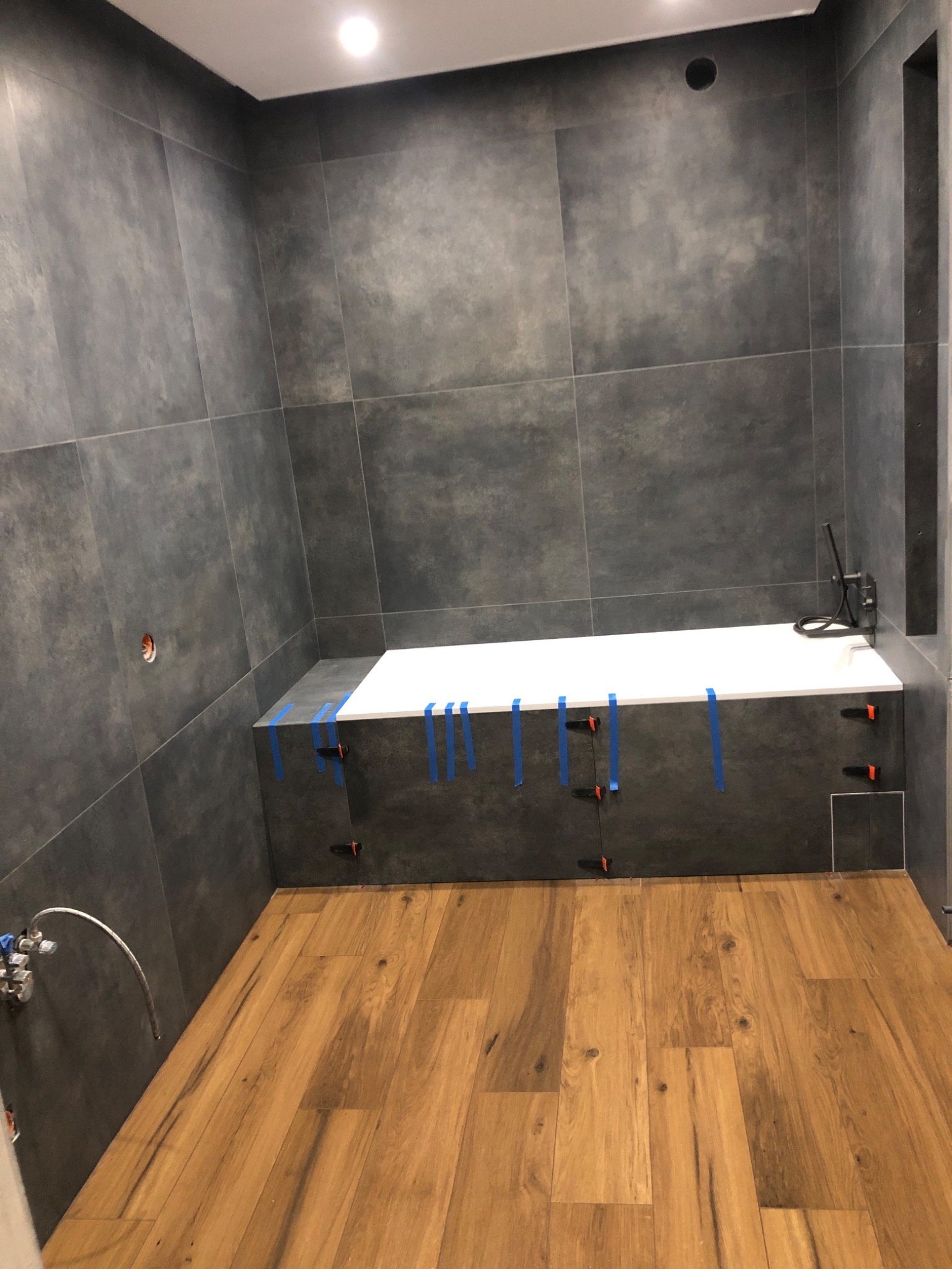 Łazienka w trakcie prac: szare płytki wielkoformatowe na ścianach, drewnopodobna podłoga, obudowa wanny z białą wanną zabezpieczona niebieską taśmą i pomarańczowymi krzyżykami dystansowymi.