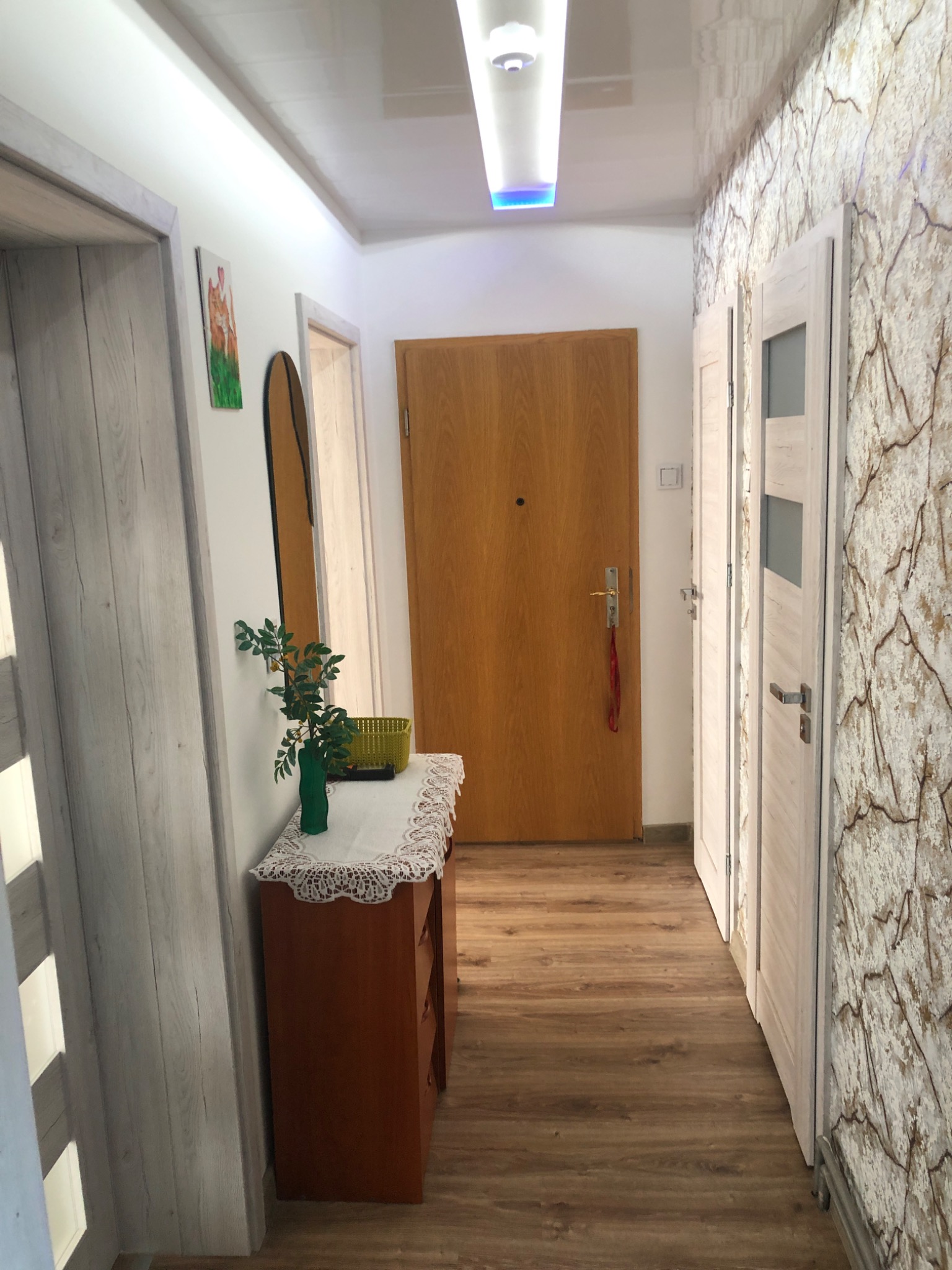 Korytarz po remoncie: drewnopodobne panele podłogowe, ściana z dekoracyjną tapetą imitującą kamień, brązowe drzwi wejściowe, białe drzwi wewnętrzne z przeszkleniami, błyszczący sufit z oświetleniem...