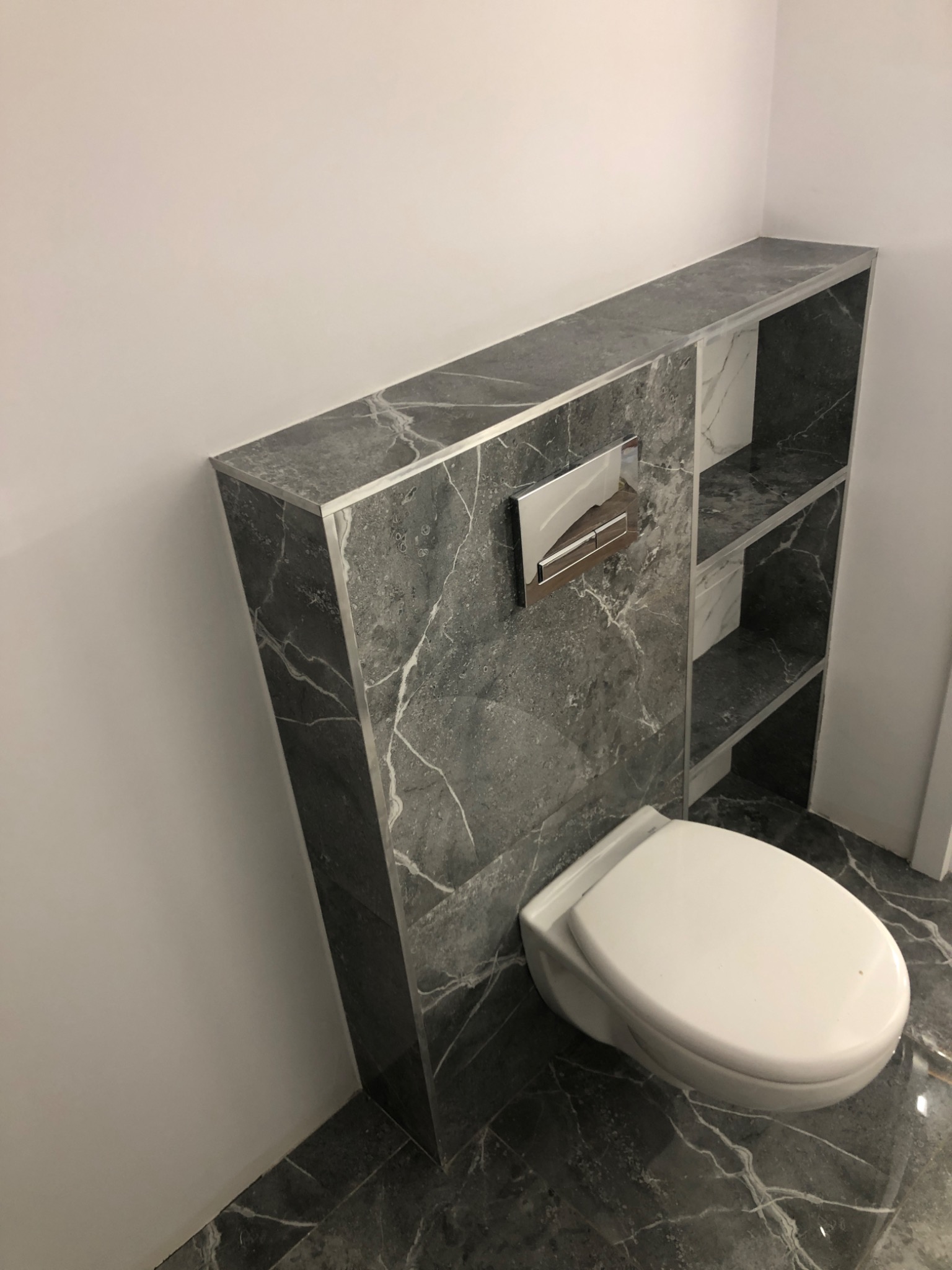 Nowoczesna toaleta podwieszana z szarym marmurowym wykończeniem, wnęki półek nad spłuczką, chromowany przycisk spłukiwania, marmurowa posadzka.