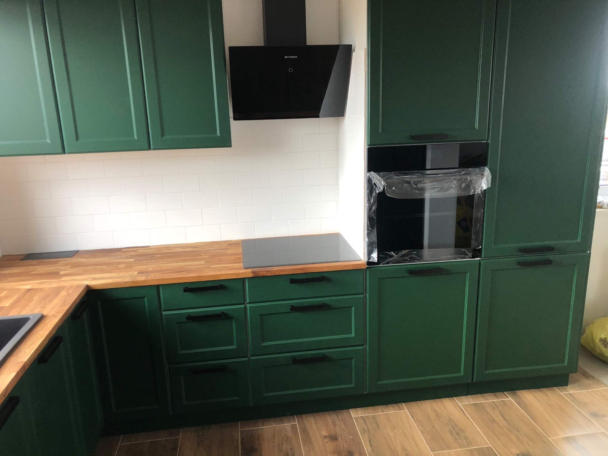 Nowoczesna kuchnia z zielonymi szafkami, drewnianym blatem, białymi płytkami na ścianie i czarnym okapem oraz piekarnikiem w folii ochronnej.