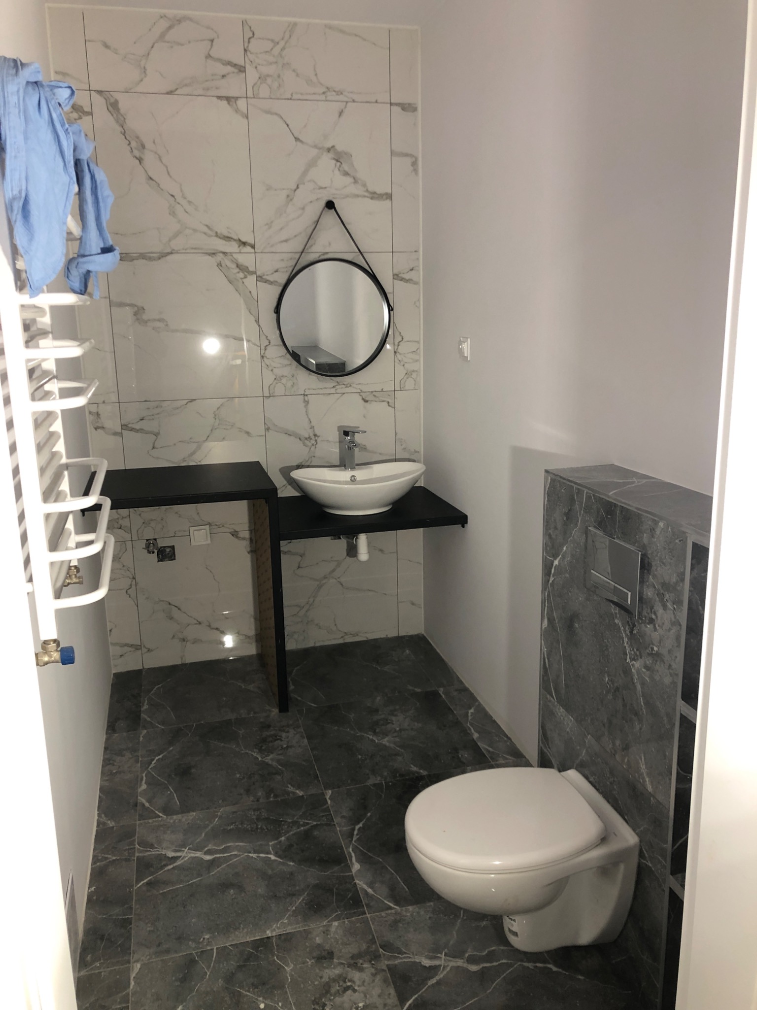 Nowoczesna łazienka z marmurowymi płytkami, wiszącą toaletą i umywalką nablatową na czarnym blacie, okrągłe lustro na ścianie, grzejnik drabinkowy z niebieskim ręcznikiem.