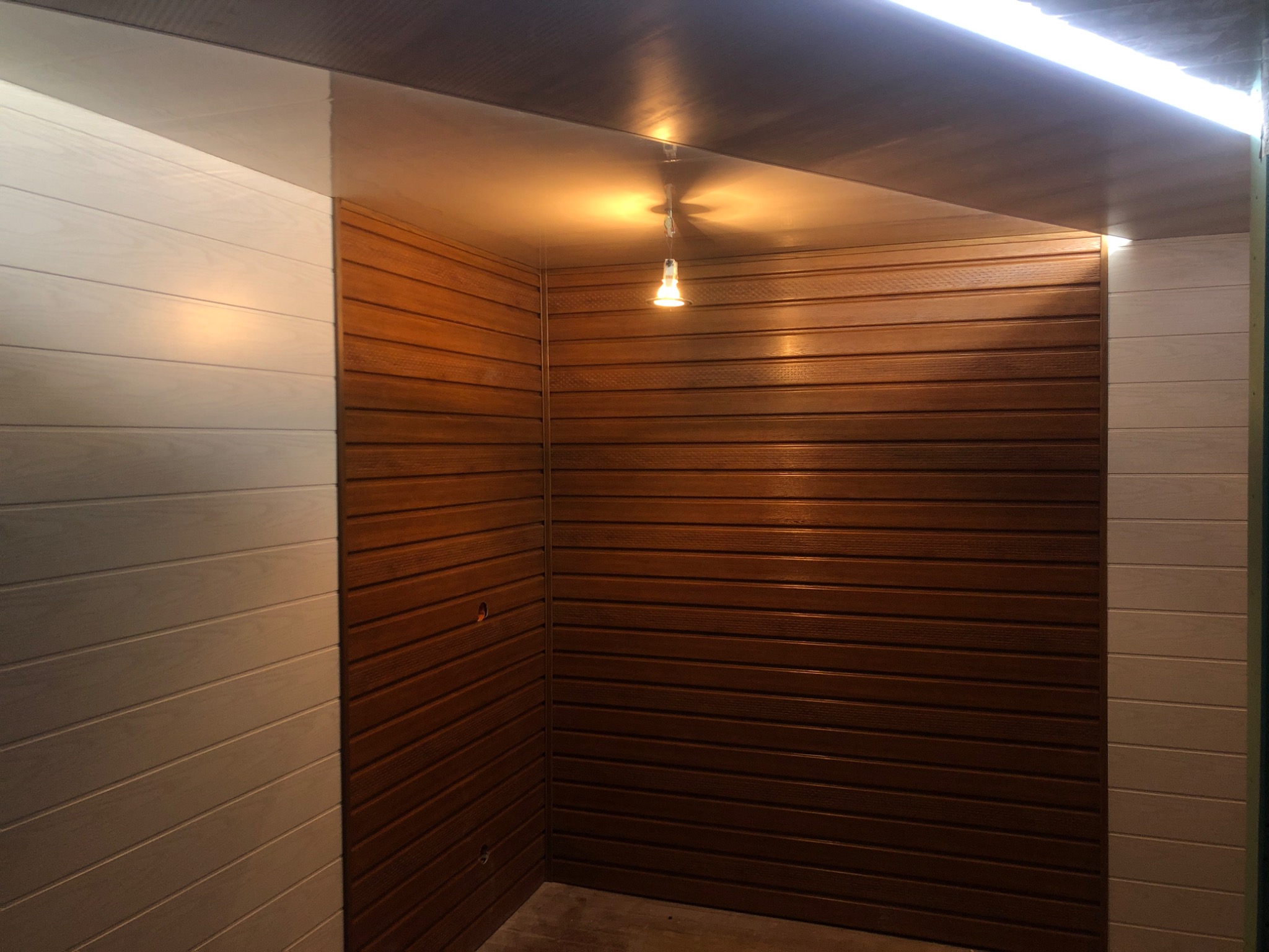 Wnętrze wykończone panelami ściennymi w dwóch kolorach: brązowym i kremowym, oświetlone wiszącą żarówką i listwą LED na suficie.