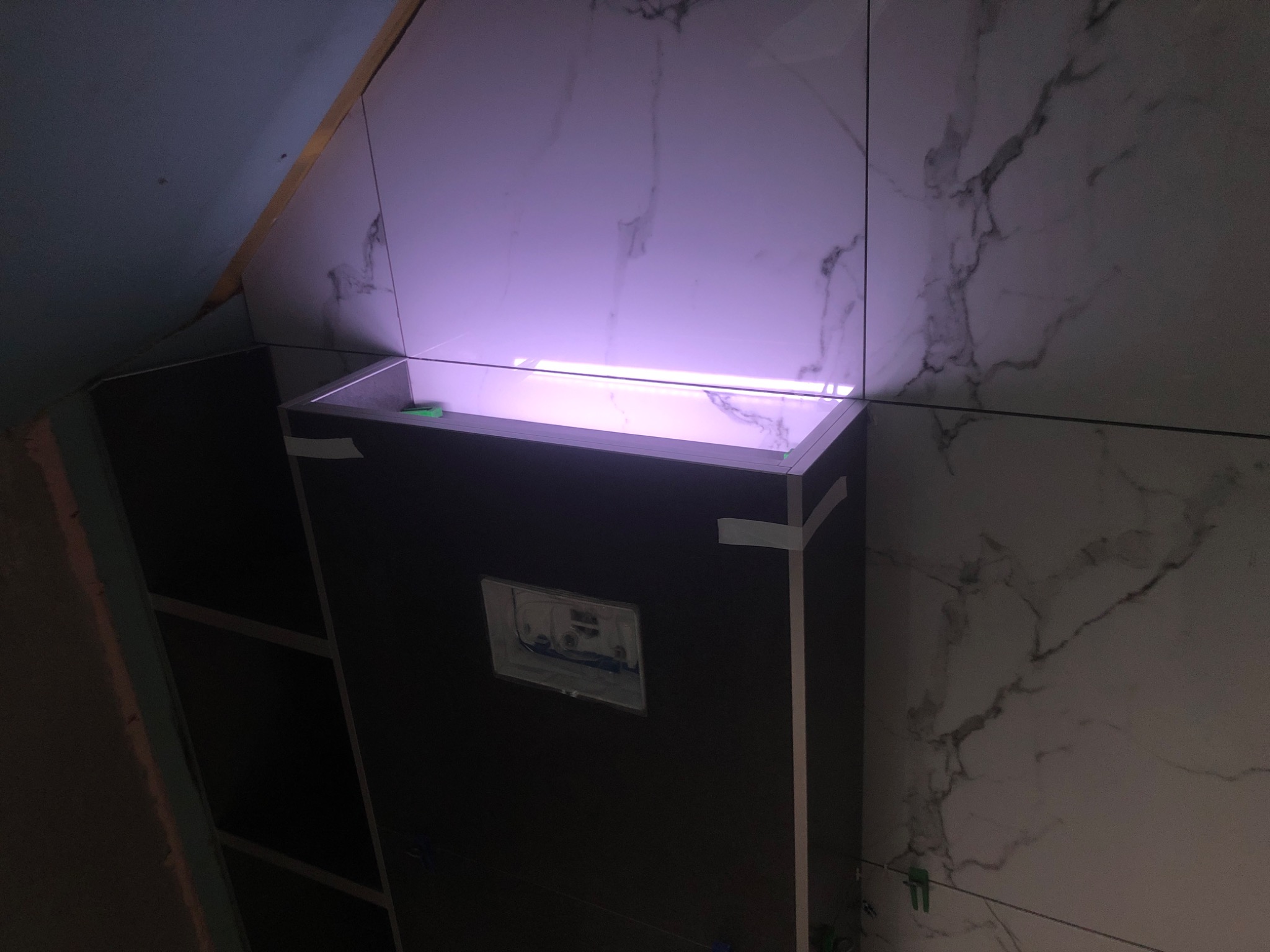 Stelaż podtynkowy WC obudowany płytami, podświetlony od góry, z fragmentem ściany wykończonej płytkami imitującymi marmur, widoczne taśmy maskujące i elementy poziomowania płytek.