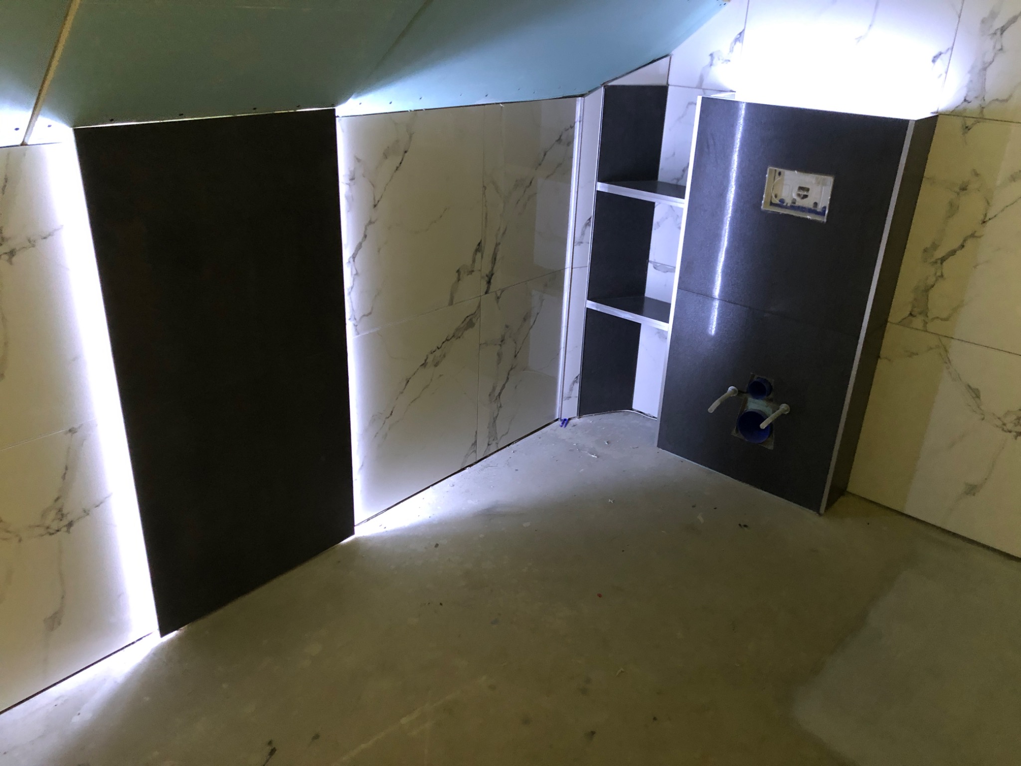 Częściowo wykończona łazienka z czarno-białymi płytkami imitującymi marmur, widoczne instalacje wodne i otwarta półka w zabudowie.