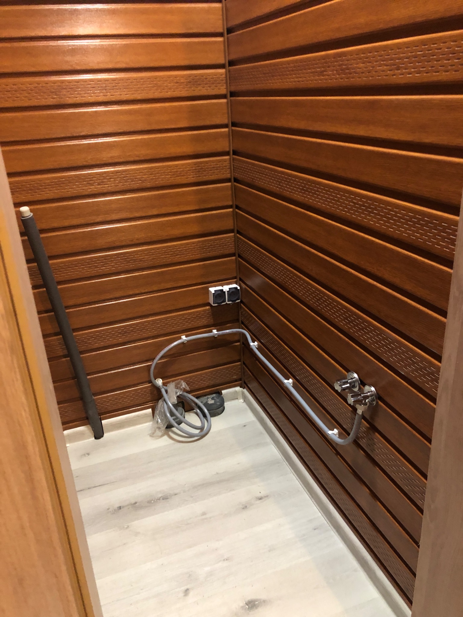 Wnętrze małej łazienki wykończonej panelami ściennymi w kolorze ciemnego drewna, widoczna instalacja wodna z baterią i rurami, podłoga wyłożona jasnymi panelami.