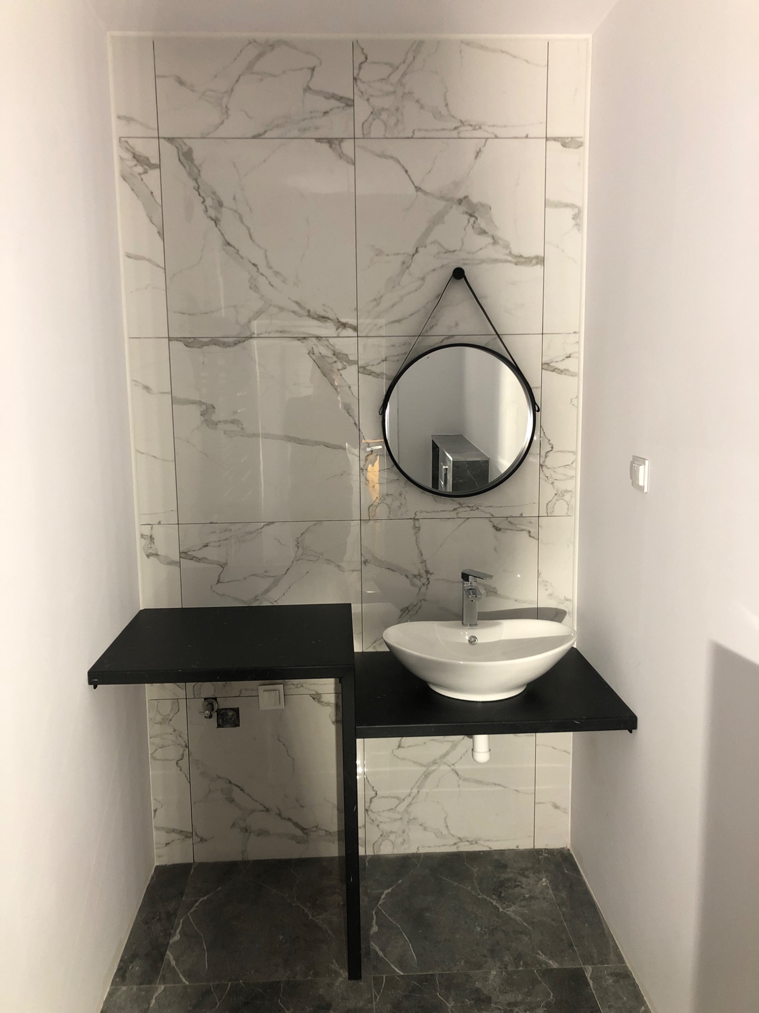 Nowoczesna łazienka z umywalką nablatową, okrągłym lustrem na tle białych płytek imitujących marmur oraz czarnym blatem i stelażem.