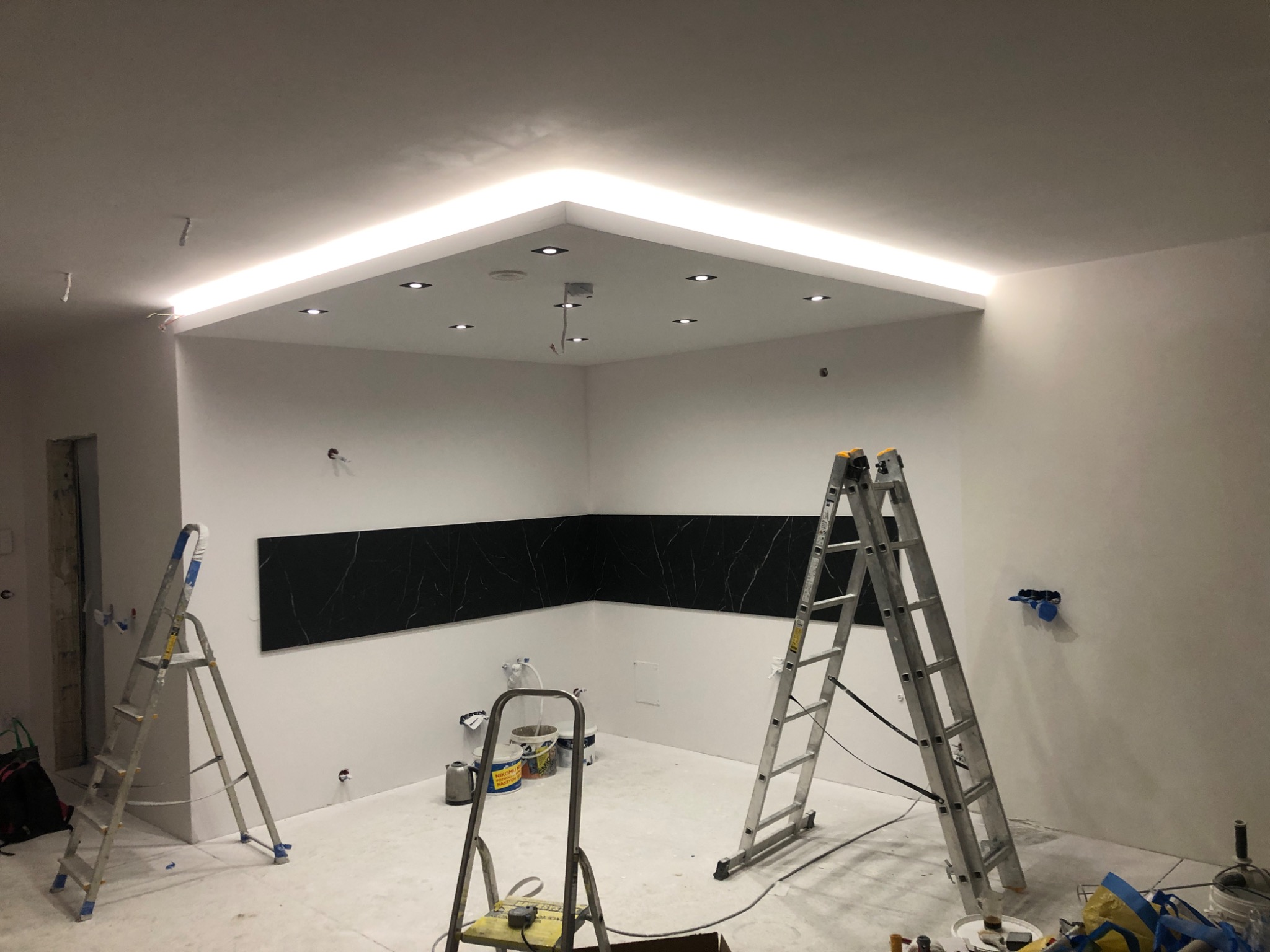 Wnętrze w trakcie prac wykończeniowych: czarne panele ścienne imitujące marmur, oświetlenie sufitowe LED, dwie drabiny aluminiowe, ściany przygotowane do malowania, widoczne puszki z farbą...
