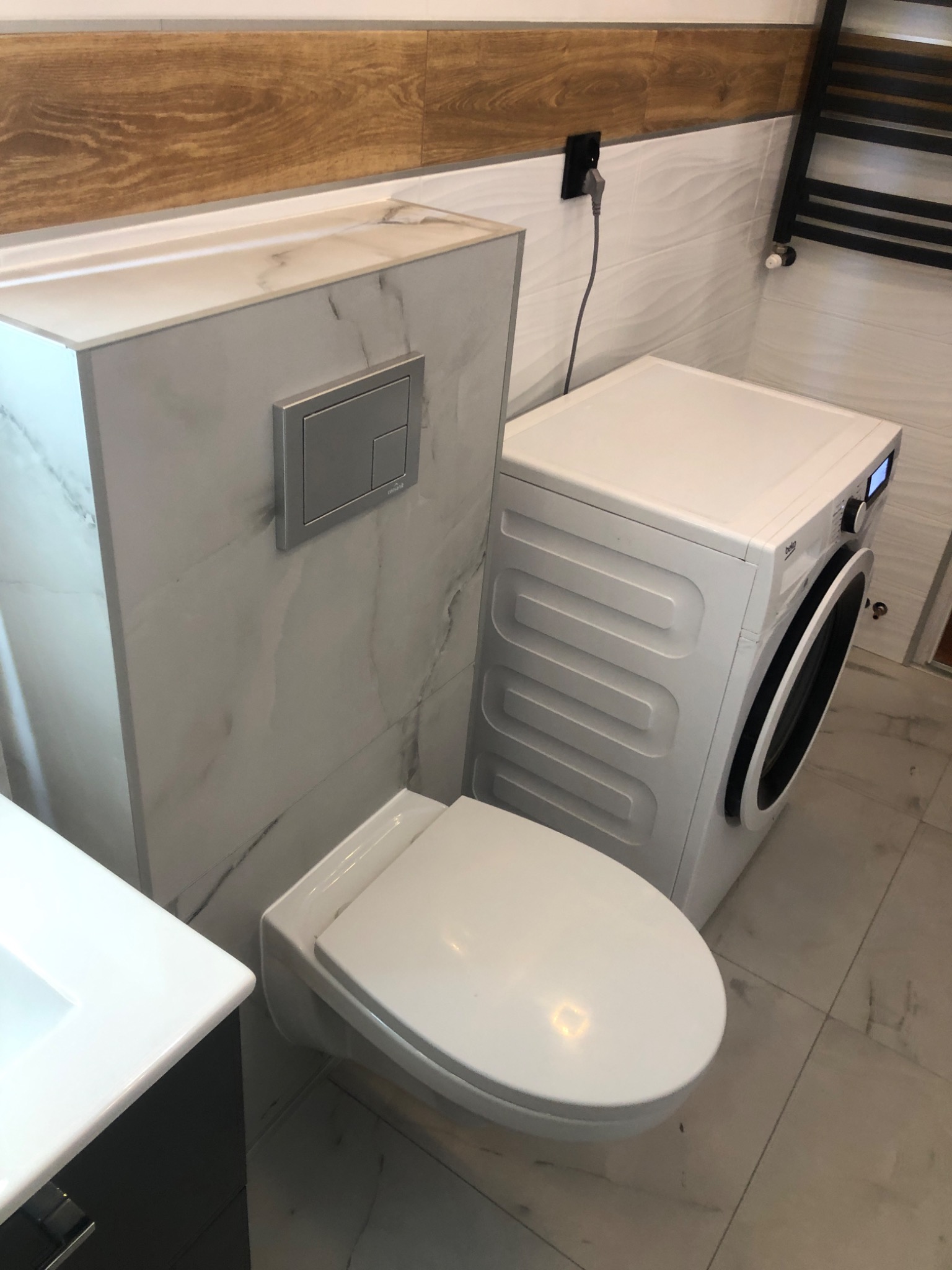 Nowoczesna łazienka z podwieszaną miską WC obudowaną płytą imitującą marmur, pralką Beko i grzejnikiem drabinkowym na ścianie wykończonej płytkami z delikatnym wzorem fal.