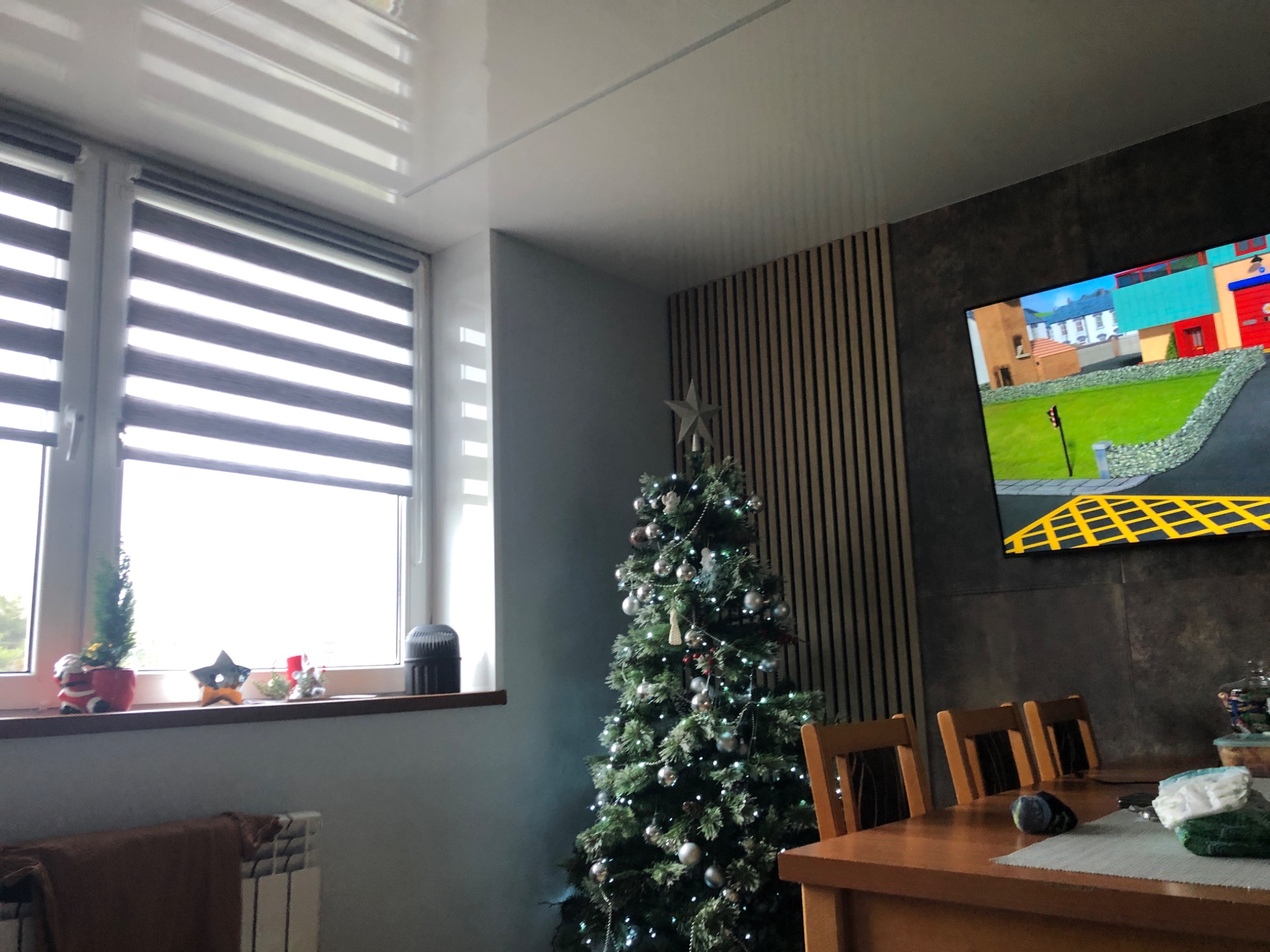 Salon z błyszczącym sufitem, żaluzjami okiennymi, dekoracjami na parapecie, choinką z srebrnymi bombkami, drewnianą ścianą z pionowymi panelami i telewizorem z włączoną bajką.