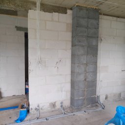 Usługi elektryczne Mikołajki - Surowy stan budynku w trakcie prac: kable elektryczne prowadzone po ścianach z bloczków betonowych, widoczne peszle i przewody, niedokończony komin z pianką montażową na szczycie.