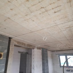 Usługi elektryczne Mikołajki - Surowy betonowy strop z widocznymi śladami szalunku i prowadzonymi przewodami elektrycznymi, ściany z bloczków betonowych i gazobetonowych w surowym stanie, widoczne przygotowania do dalszych prac...