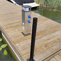 Usługi elektryczne Mikołajki - Srebrna skrzynka z gniazdami elektrycznymi na drewnianym pomoście, obok czarna rura. W tle łódź i trzciny. Instalacja elektryczna na przystani.