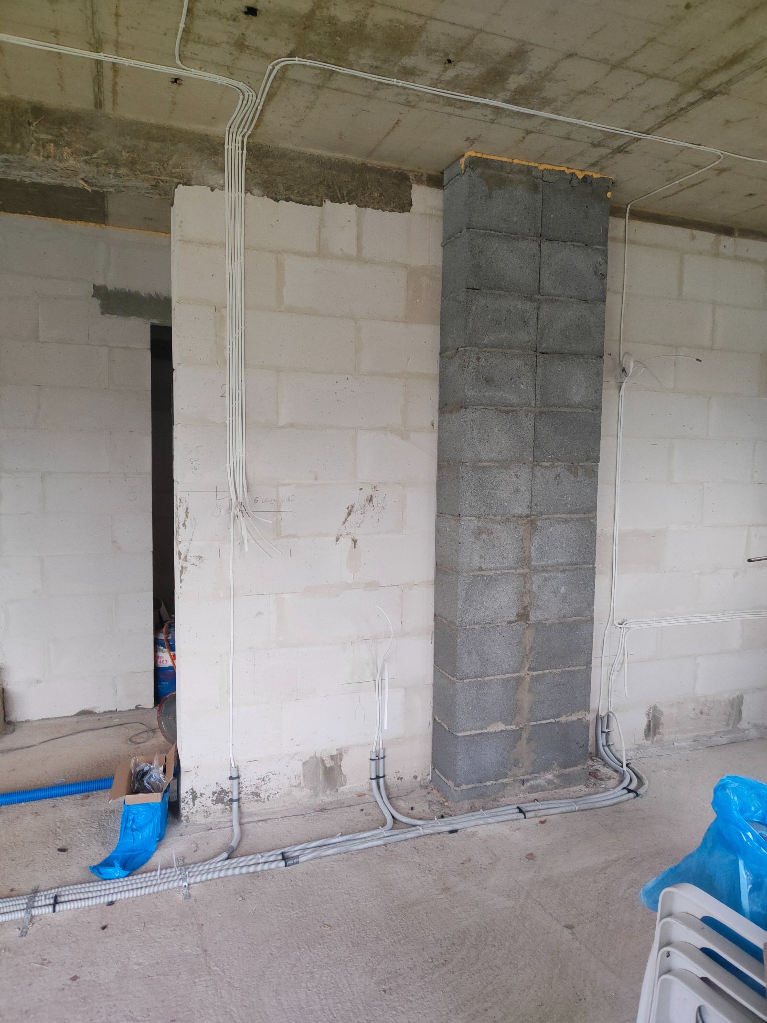 Surowy stan budynku w trakcie prac: kable elektryczne prowadzone po ścianach z bloczków betonowych, widoczne peszle i przewody, niedokończony komin z pianką montażową na szczycie.