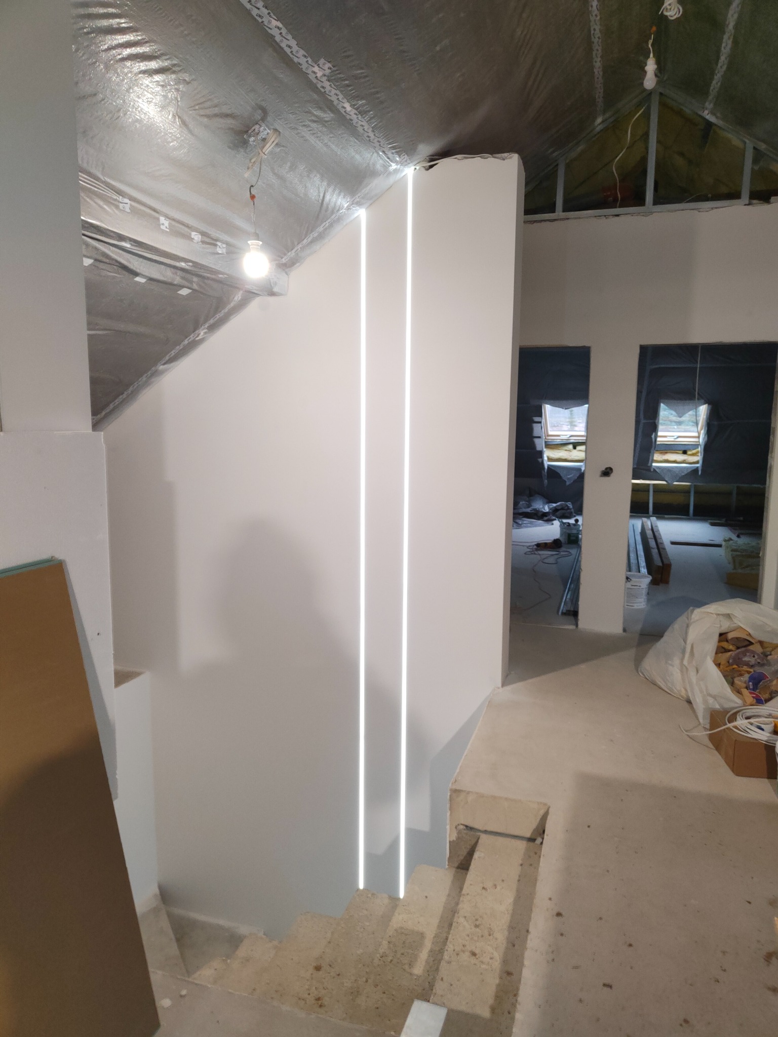 Wnętrze poddasza w trakcie wykańczania, widoczne dwie pionowe linie oświetlenia LED na białej ścianie, surowe schody i ocieplenie dachu folią aluminiową.