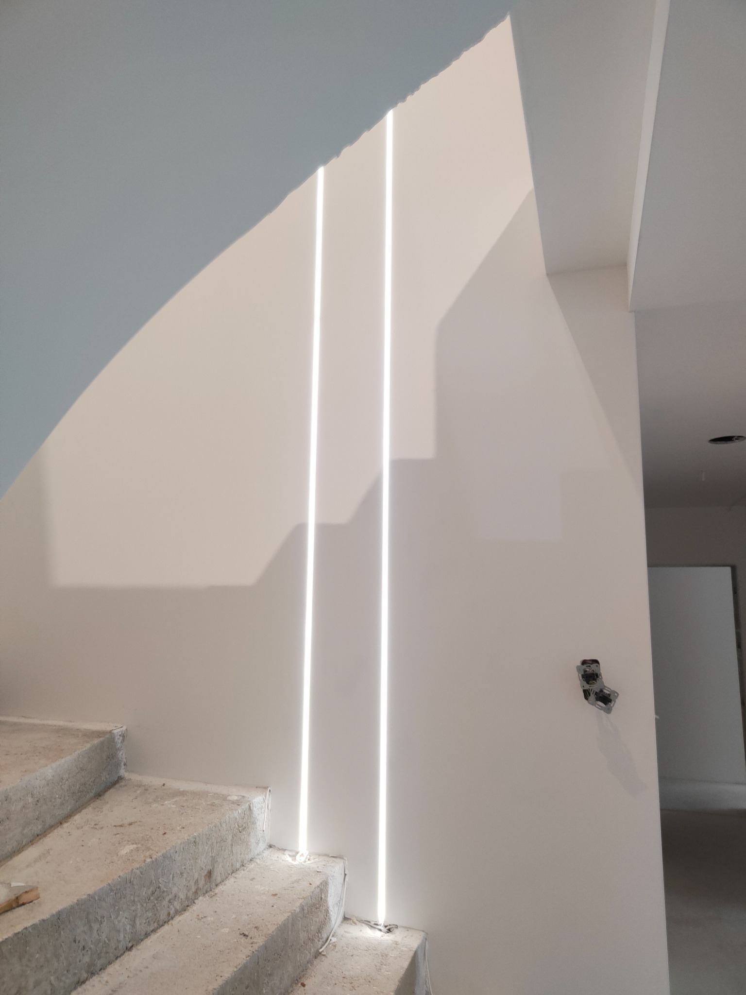 Surowe betonowe schody w surowym stanie z trzema pionowymi pasami oświetlenia LED w ścianie i gniazdkiem elektrycznym bez osprzętu.